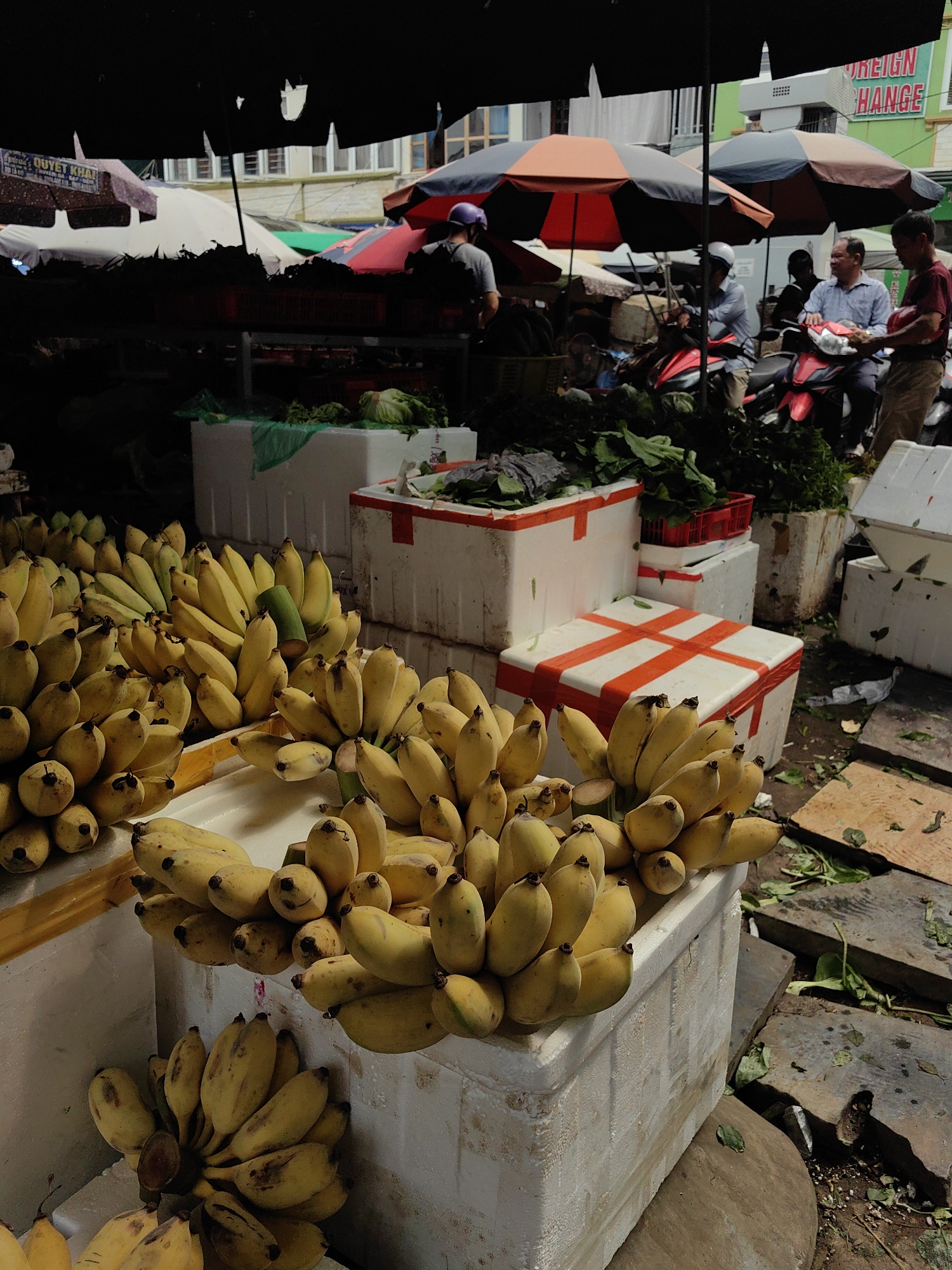 The image shows a bustling outdoor market scene, likely in Hải Phòng, Vietnam, given the types of goods being sold. The primary focus is on several crates filled with bananas of varying ripeness, ranging from green to yellow. The bananas are piled high in white, weathered polystyrene boxes, with some boxes stacked on top of others. To the right, there's a stack of wooden planks with plant debris scattered around. In the background, there are vendors under umbrellas, some on motorbikes, and others standing around. One vendor in a blue shirt is sitting on a motorbike, seemingly interacting with another person in a maroon shirt. A sign indicating "FOREIGN EXCHANGE" is visible on a green building in the background. The scene is illuminated by natural light, with some areas in shadow. Other produce is also visible in the background, adding to the market atmosphere.