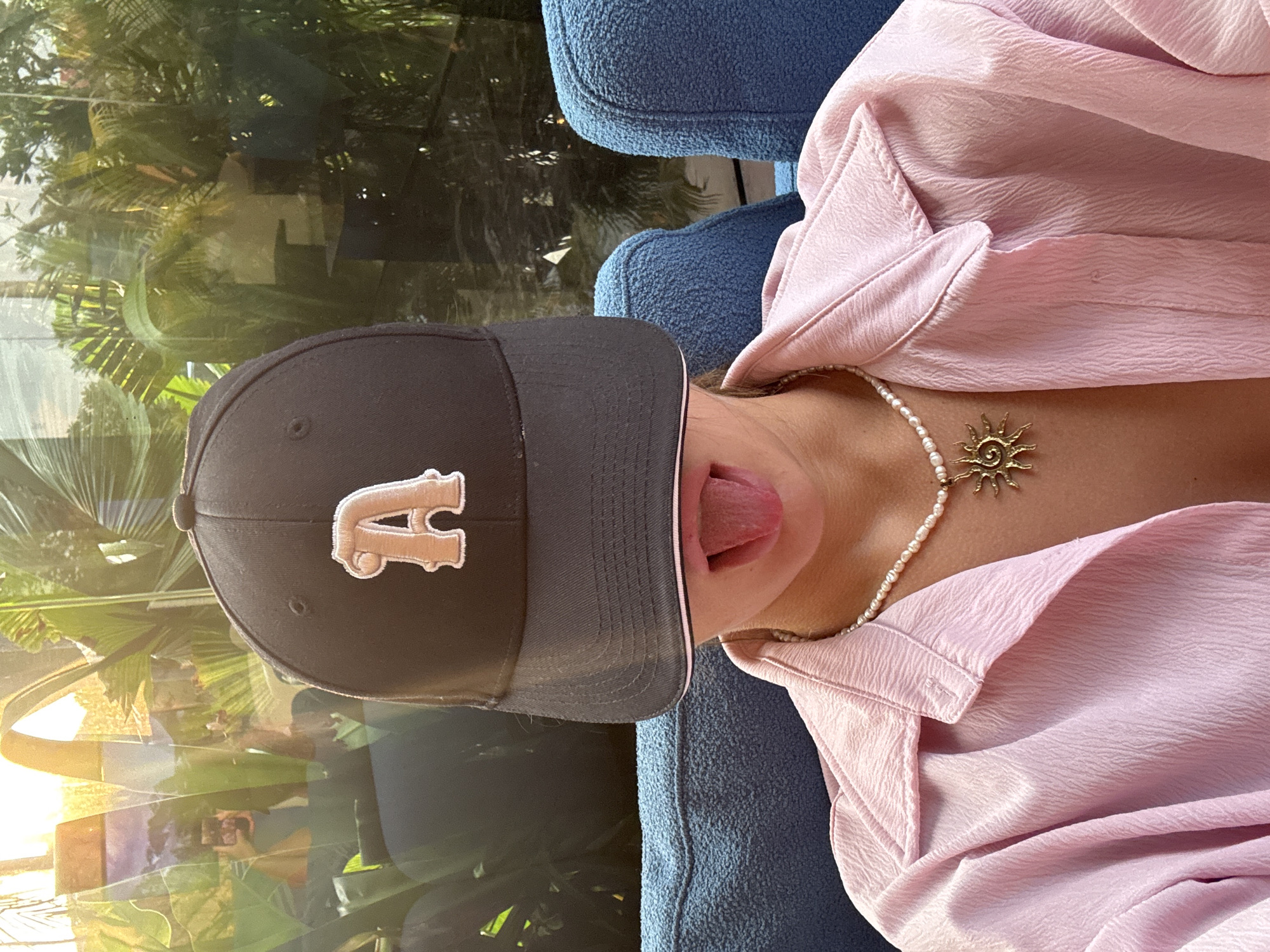 An individual is captured in a close-up, upward-angled shot, playfully sticking their tongue out. They are wearing a black baseball cap with a prominent beige or light-colored letter "A" embroidered on the front. Below the cap, a strand of pearls forms a necklace, adorned with a bronze-toned, sun-shaped pendant featuring radiating rays and a central face design. The person is also wearing a light pink, textured shirt or top with visible folds and wrinkles.
The individual appears to be seated on a blue, textured armchair or couch, with its fabric visible around their neck and arm area. The background features a large glass partition or window, reflecting the surroundings. Through the glass and in its reflections, lush green tropical foliage, resembling palm fronds and other dense vegetation, is clearly visible. The reflection in the glass also reveals the individual holding a smartphone, indicating they are taking a selfie. Sunlight streams in, suggesting a bright daytime setting. The overall scene implies a relaxed, outdoor or semi-outdoor setting in a tropical location, consistent with North Kuta, Indonesia.