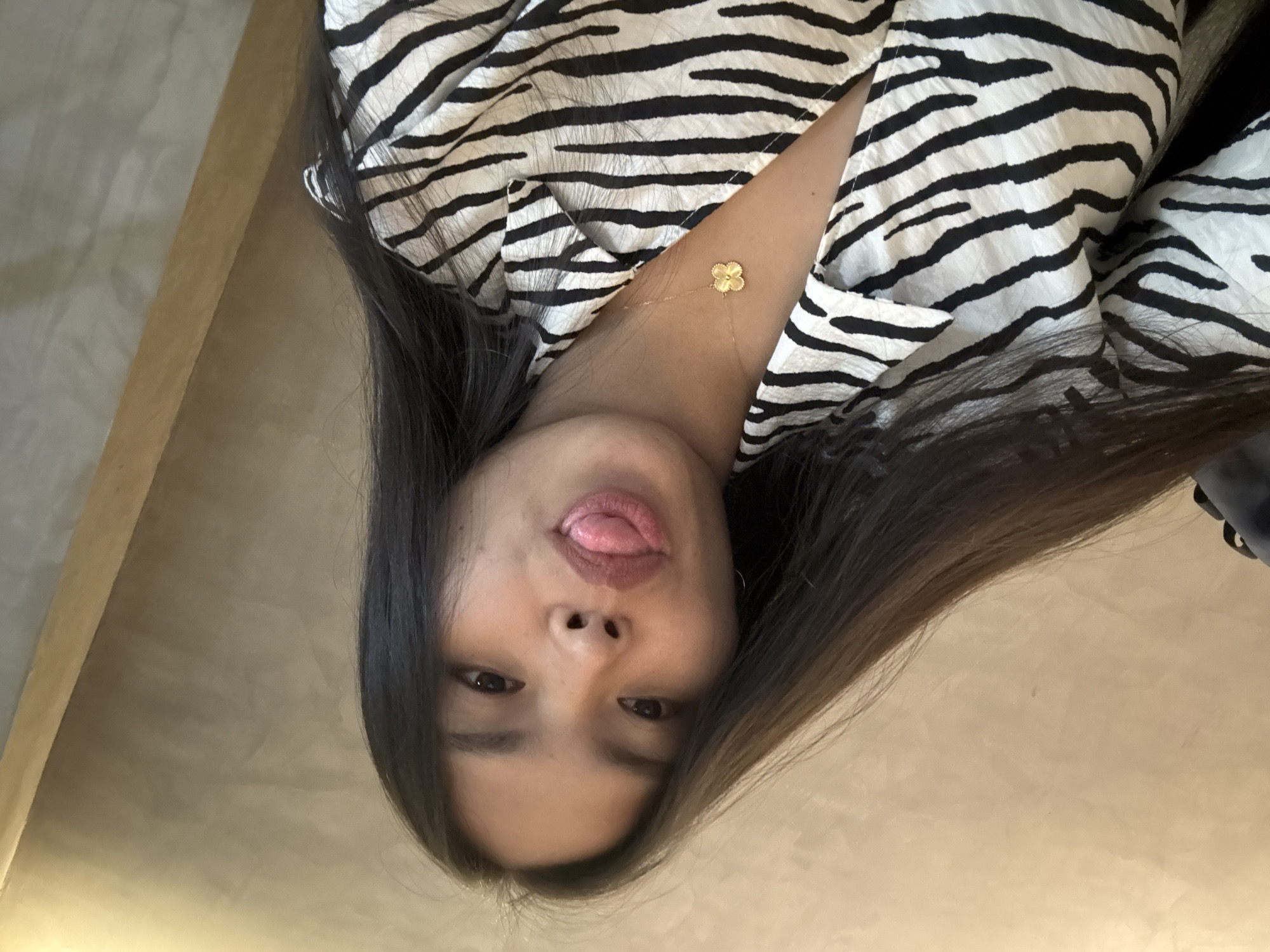 A young adult female with dark hair is visible in the image. She is wearing a zebra-striped shirt and a gold necklace with a four-leaf clover pendant. She is sticking her tongue out. The background is out of focus. There is a light-colored wall or surface. The location is unknown and cannot be determined from the image. The image does not reveal any information indicating the city of Davao City, Philippines.