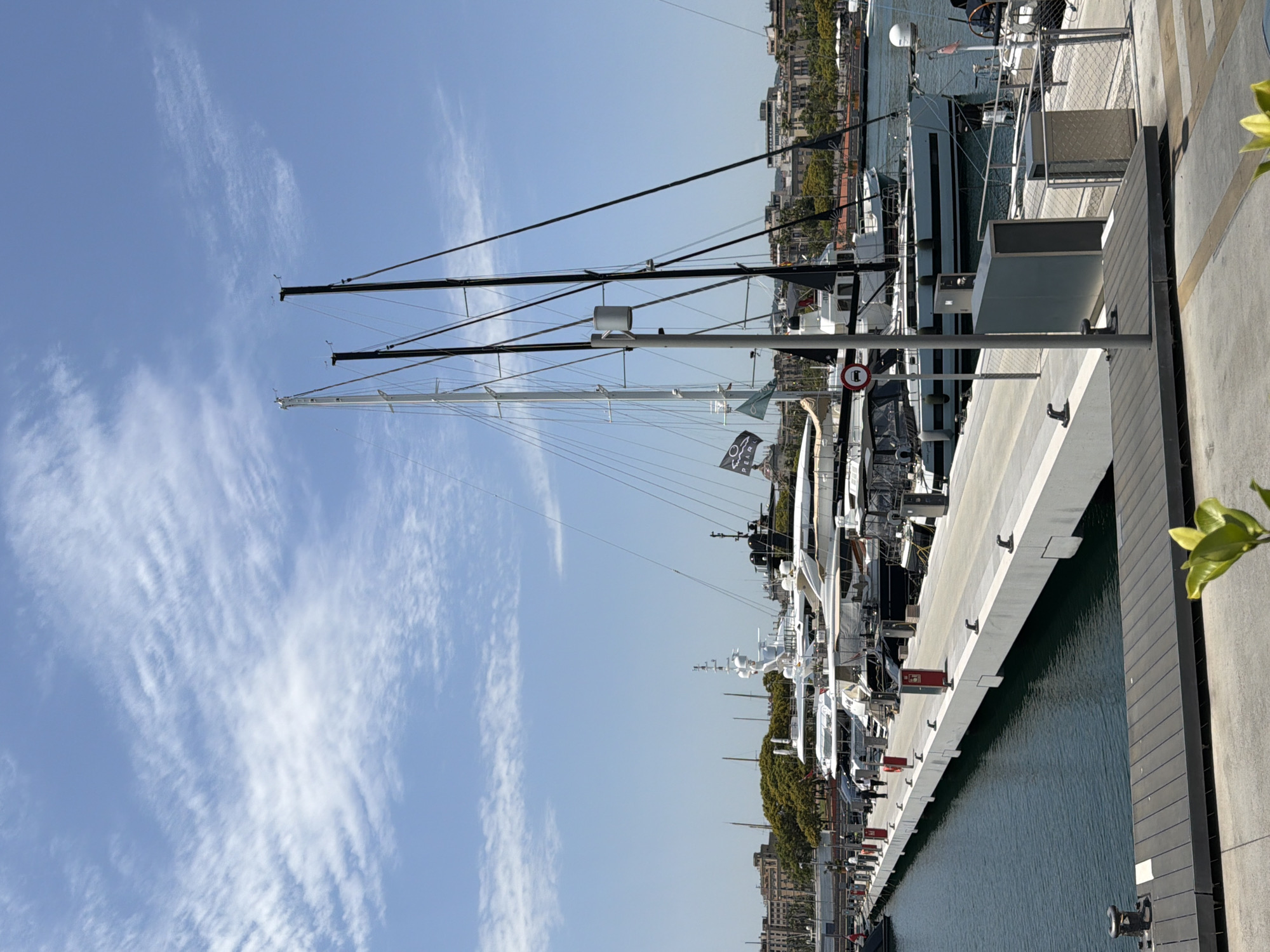 The image depicts a marina in Barcelona, Spain, filled with numerous yachts docked along a pier. The sky is a clear blue with scattered wispy clouds, suggesting fair weather. Sunlight casts shadows, indicating it is likely daytime, possibly late morning or early afternoon.
The foreground features a concrete pier with dark wooden planks along its edge, a metal bollard, and a few yellow leaves from an unseen plant. Several yachts are moored in the water, their masts and rigging extending high into the sky. The yachts are of various sizes, with some being sleek and modern, while others appear more traditional. One yacht prominently displays a black flag with white text that reads "PEARL." Further in the background, a cityscape with buildings and lush green trees lines the waterfront.
The scene is tranquil, with no people clearly visible, suggesting a quiet moment in the bustling marina. The overall impression is one of luxury and leisure, typical of a high-end yachting destination. The image is oriented vertically, with the sky occupying the upper portion and the marina and city in the lower portion.