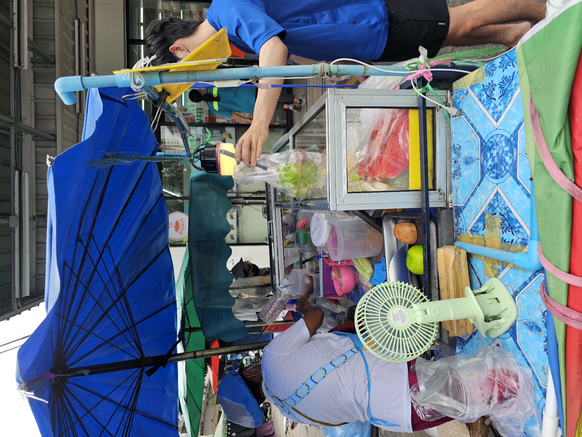 The image depicts an outdoor food stall in Bangkok, Thailand. A vendor wearing a white shirt, blue apron, and a blue hairnet is preparing food behind a glass display counter. A customer, a young man in a blue t-shirt and black shorts, is reaching into the counter to take a bag of produce. The stall is covered by a large blue umbrella and features various food items, plastic containers, a small green fan, and a yellow flag. In the background, other market stalls and shops are visible, along with pedestrians. The scene suggests a bustling local market environment.