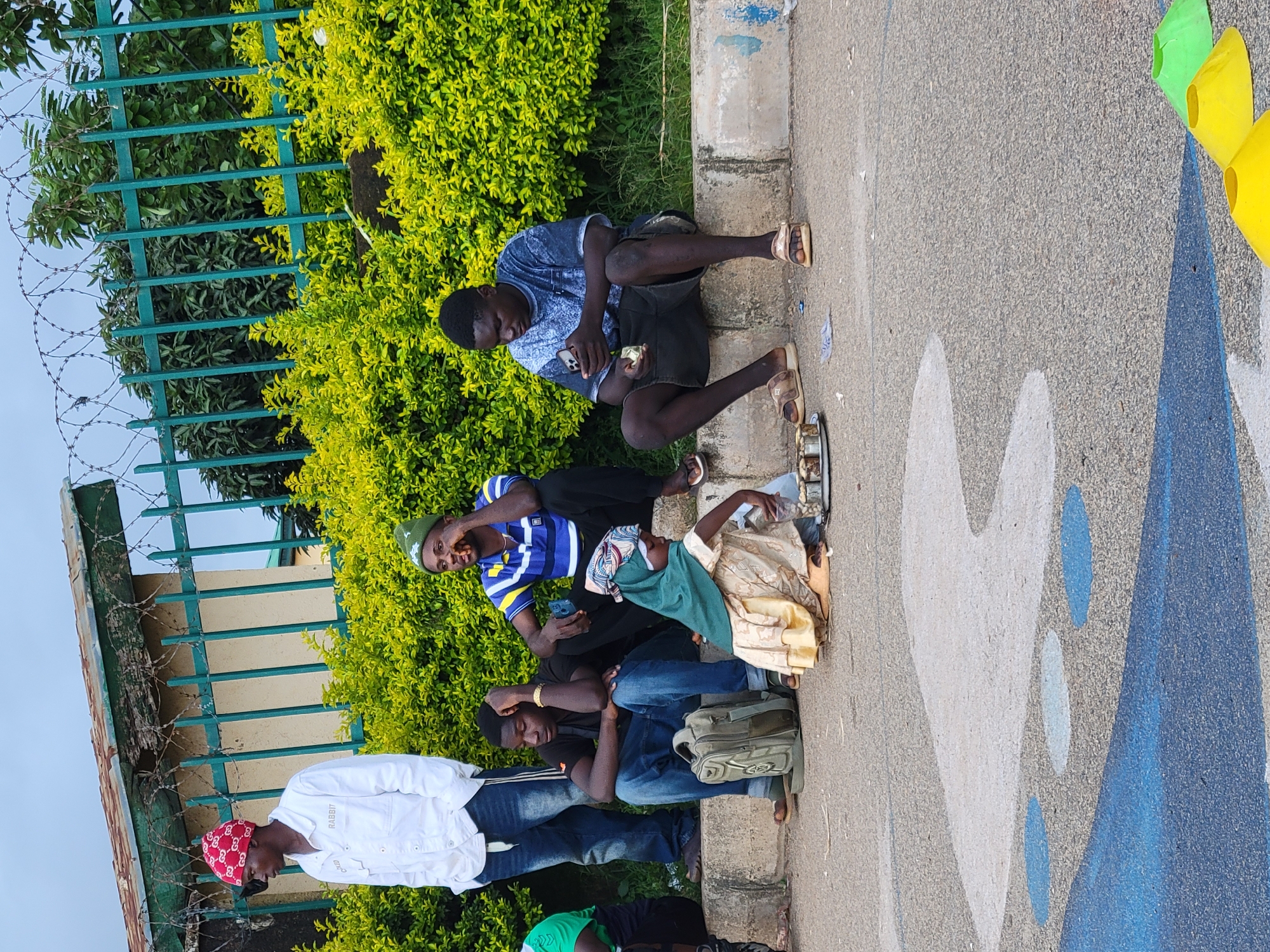 In Jos, Nigeria, a street scene unfolds with a group of individuals gathered on a concrete curb. The foreground is a paved surface with abstract blue and grey painted markings, and several yellow and green traffic cones are visible in the lower right corner.
Behind the curb, a verdant hedge forms a lush backdrop. A green metal fence, topped with barbed wire, runs along the top of the hedge, separating the scene from a building with a weathered, rust-colored roof.
To the left, a young man stands, wearing a white button-down shirt and blue jeans, his left hand in his pocket and his right arm resting on the wall. He is wearing a red bandana with a white pattern on his head. Next to him, sitting on the curb, is another young man in a black t-shirt and blue jeans, his left arm resting on his knee and his head tilted down. A green backpack rests beside him.
To the right of the seated young man, another individual in a blue and white striped t-shirt and black pants sits on the curb, holding what appears to be food up to his mouth. Further to the right, a young man in a blue tie-dye t-shirt and black shorts sits on the curb, looking down at his mobile phone. He is wearing sandals.
In front of the seated individuals, a young girl in a light-colored dress and a green headscarf is kneeling on the ground. She is attending to a tray of small items, possibly food or goods for sale, arranged on a metal plate. She is holding a plastic bag.
The sky above is overcast, suggesting a cloudy day. The overall impression is a casual gathering of people on a street in Jos, possibly waiting or socializing.