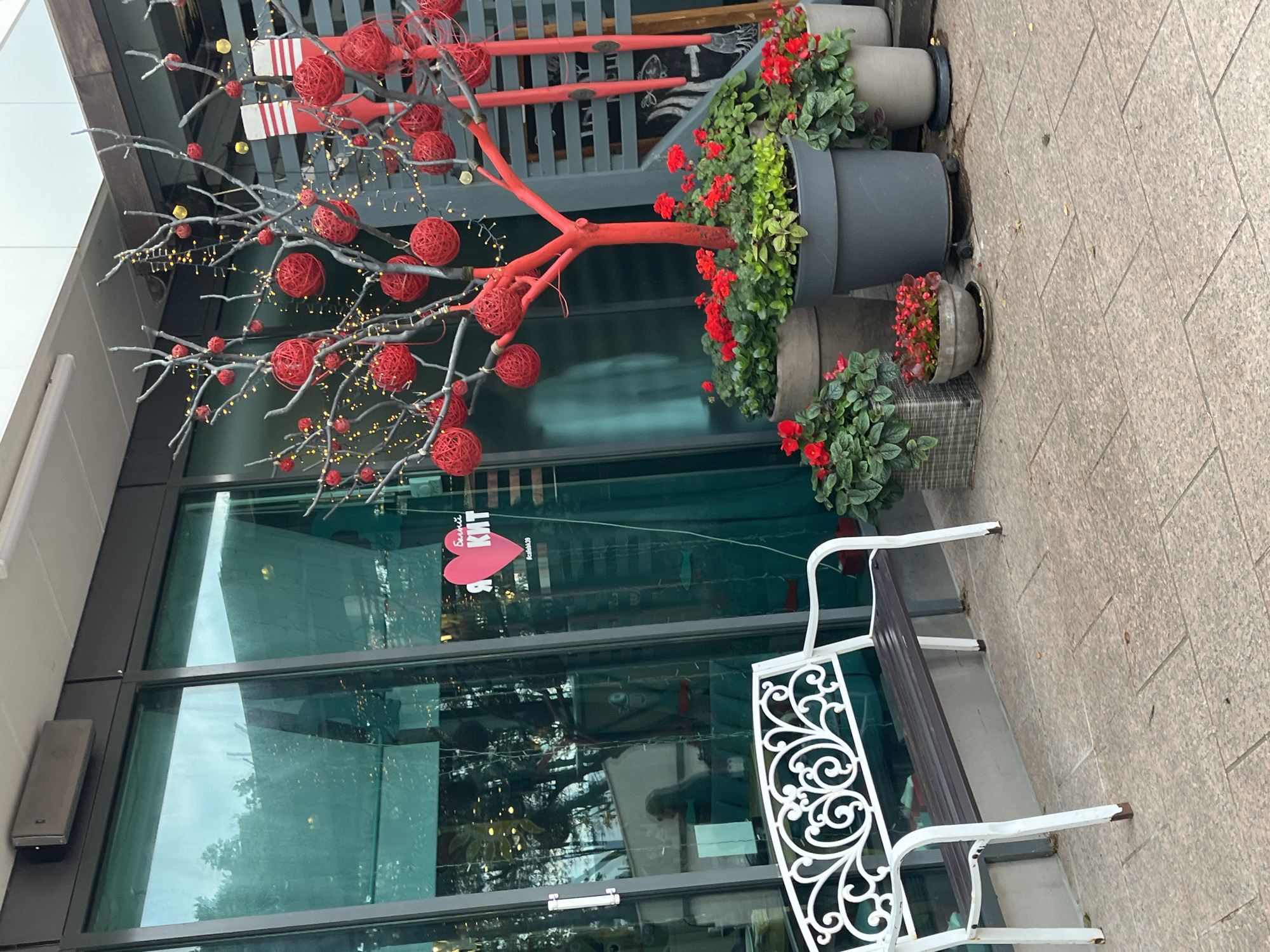 A white metal bench with a dark brown wooden seat sits on a tiled patio in front of a building with large glass windows. To the right of the bench is a decorative red tree adorned with red ball ornaments and small yellow lights. In front of the tree are several planters filled with red flowers and green foliage. A pink heart-shaped sticker with Russian text, including "БЕЛЫЙ КИТ" (White Whale) and "#cafebk39", is visible on the glass window. The scene appears to be an outdoor seating area for a cafe or shop in Svetlogorsk, Russia.