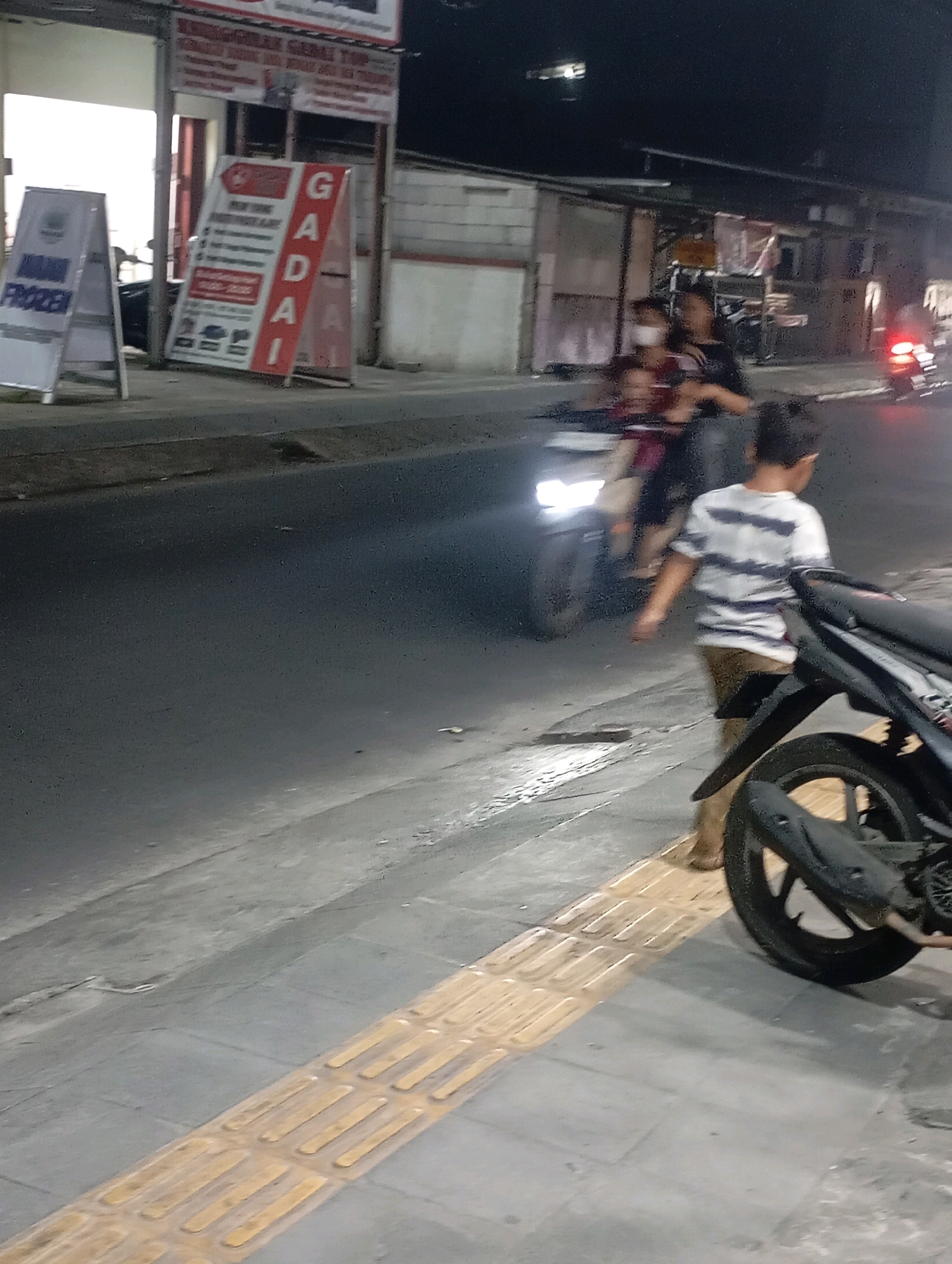 The image depicts a street scene at night in Babelan, Indonesia. In the foreground, a child wearing a striped t-shirt and shorts stands on a tactile paving surface. Next to the child, the rear wheel and exhaust of a motorcycle are visible. In the middle ground, a group of people are on a motorcycle, with one person appearing to be driving and two passengers, one of whom is a child. Headlights from the motorcycle are illuminated. On the left side of the image, A-frame signs are displayed, one with the word "FROZEN" and another with red and white lettering, including the word "GADAI," which translates to "pawnshop." The background shows a building facade with a light emanating from an open doorway. The street surface appears wet, reflecting the ambient light.