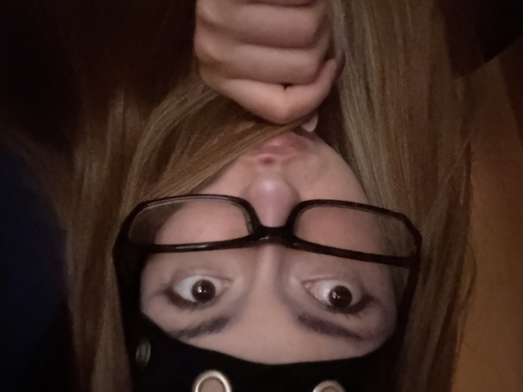 An upside-down selfie of a person wearing glasses and a black face mask with metal grommets. The person's hair is blonde and is held above their face by a hand. The location is unknown.