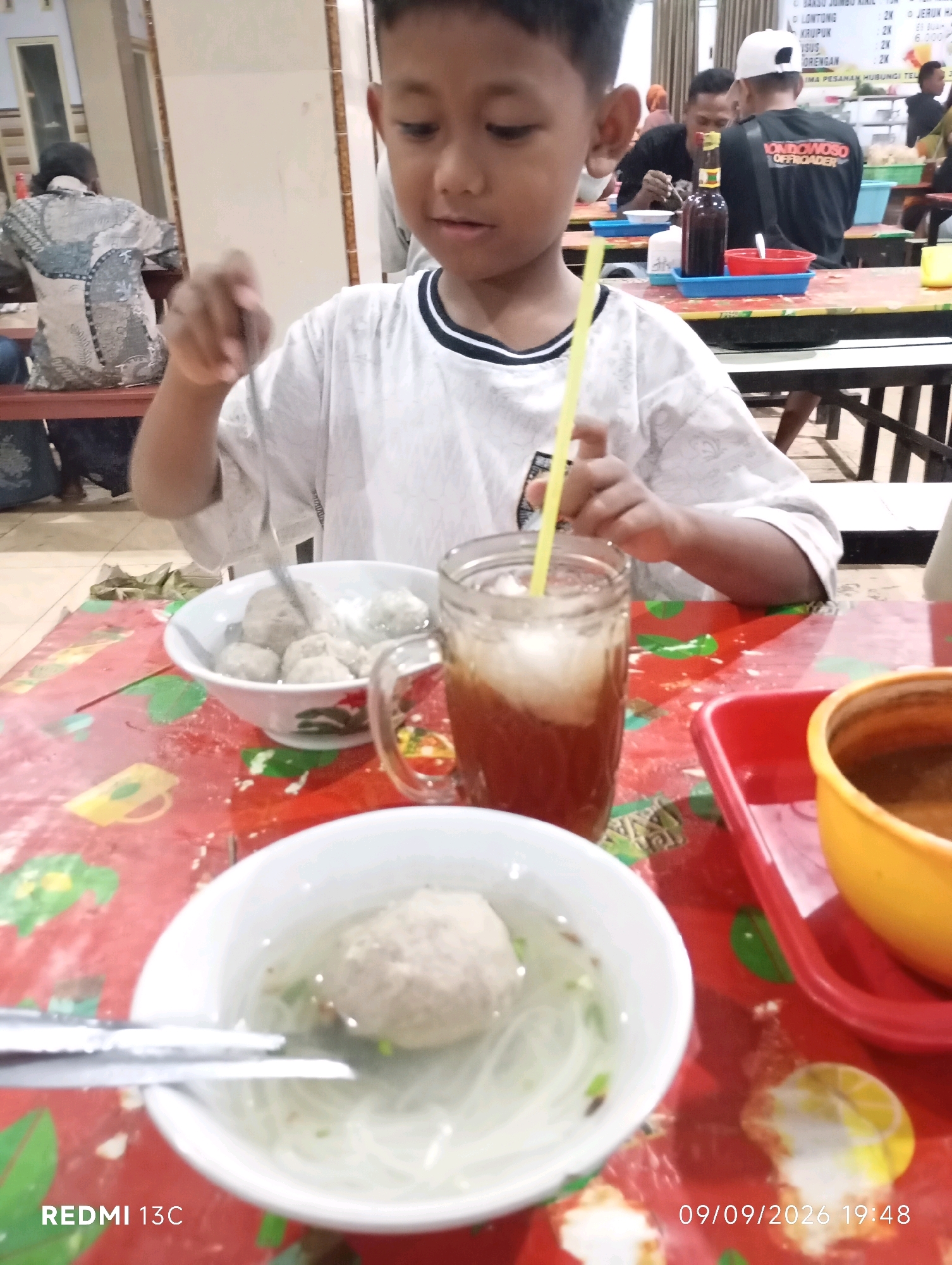 A young boy with dark hair and a white t-shirt is seated at a red table in an indoor dining establishment, possibly a restaurant or food stall in Wuluhan, Indonesia. He is looking down towards a bowl of food in front of him. His right hand is raised, holding a yellow straw, and his left hand is near another bowl of food. On the table are two white bowls containing meatballs and noodles, a glass of iced tea, a red plastic tray with a yellow bowl, and other items. The table surface has a colorful pattern. In the background, other patrons are visible, some seated at tables, and there are shelves with items. The environment appears to be a casual dining area.