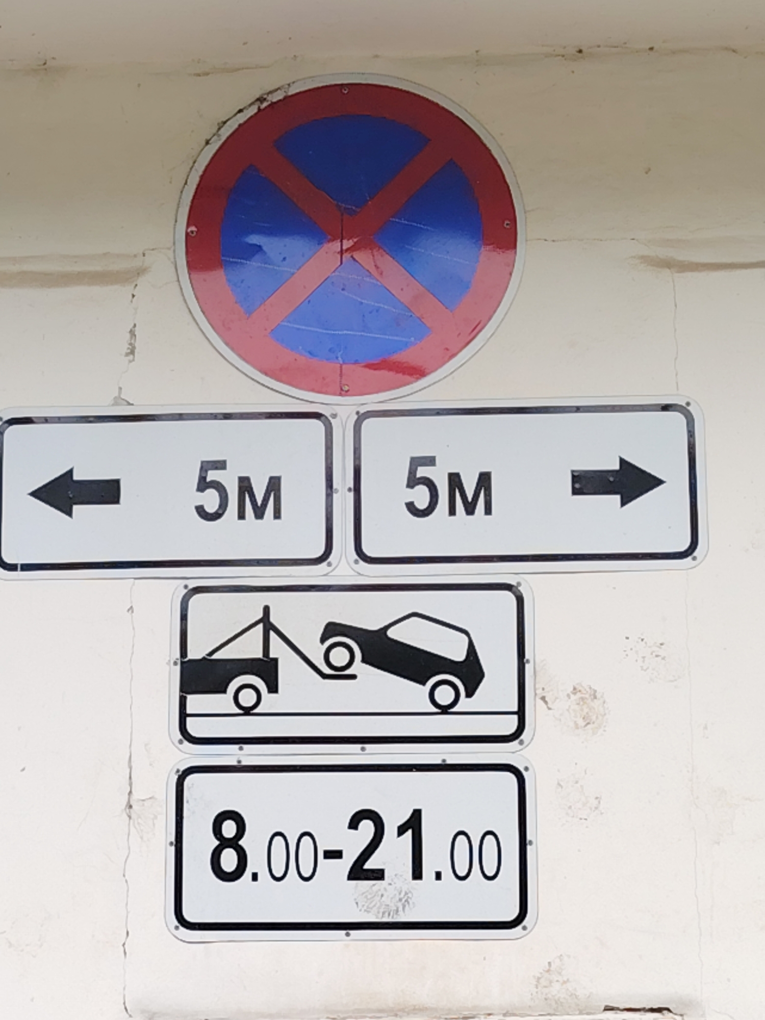 A cluster of five road signs is prominently displayed on a light-colored, somewhat weathered wall in Chaikovsky, Russia. The top sign is a standard circular "No Stopping" or "No Parking" sign, featuring a blue circle with a red border and a red diagonal cross.
Below it, two rectangular white signs provide specific distance information. The left sign shows a black left arrow followed by "5M," indicating a restricted zone extending 5 meters to the left. The right sign mirrors this, displaying "5M" followed by a black right arrow, signifying a 5-meter restricted zone to the right. Together, these suggest a 10-meter restricted stretch.
Beneath these, another rectangular white sign features a black graphic of a tow truck actively towing a car, signaling that vehicles found in violation of the restriction will be impounded. The bottom-most sign, also rectangular and white, specifies the enforcement hours with the text "8.00-21.00," indicating that the restrictions are active from 8:00 AM to 9:00 PM daily.
The wall itself shows minor wear and some staining, suggesting an outdoor urban or suburban setting. The overall scene conveys clear directives for vehicle operators regarding stopping and parking regulations.