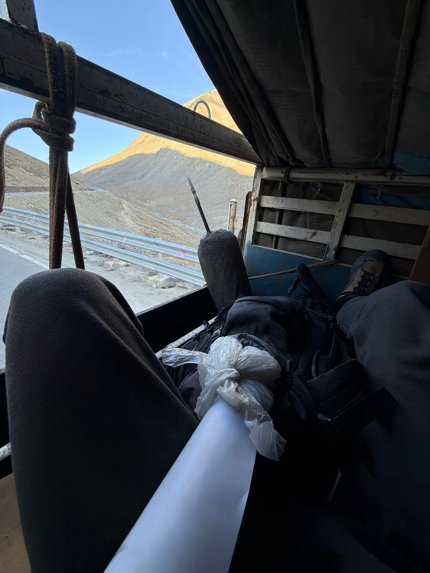 The image shows a person's legs and feet inside what appears to be the back of a vehicle, possibly a truck. The person is wearing dark pants and hiking boots. Luggage, including a backpack, is visible. A white plastic bag and a roll of white paper are also present. Outside the vehicle, a mountainous landscape with a road and guardrail can be seen. The sky is clear and blue. The overall impression is of travel in a remote, mountainous region. The scene is not identifiable as Zanskar Sumdo, India.