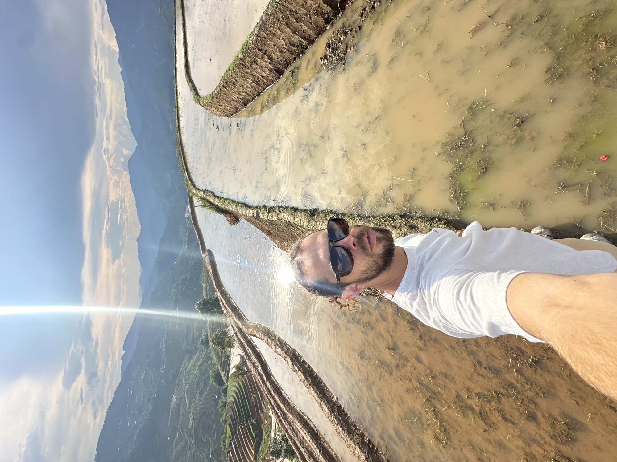 A man is captured in a selfie amidst the terraced rice paddies of Lao Chai, Vietnam. He is positioned in the foreground, facing the camera with a slight upward gaze. He has a light beard or stubble, curly hair, and wears dark sunglasses and a white t-shirt. His left arm, visible in the lower part of the frame, holds the camera for the selfie.
The man stands next to a flooded, muddy rice paddy. Extending behind him and into the distance, numerous layers of terraced rice paddies ascend the steep mountain slopes. These paddies are filled with murky brown water, some containing small green sprouts.
In the background, majestic mountains rise, partially obscured by the soft blue sky with scattered white clouds. The sun is bright, creating a prominent lens flare on the left side of the image and reflecting intensely on the water's surface. A small red cap can be seen floating in one of the water-filled paddies. The scene depicts a sunny day, likely late morning or early afternoon, showcasing the vast and beautiful agricultural landscape. No text is visible in the image.