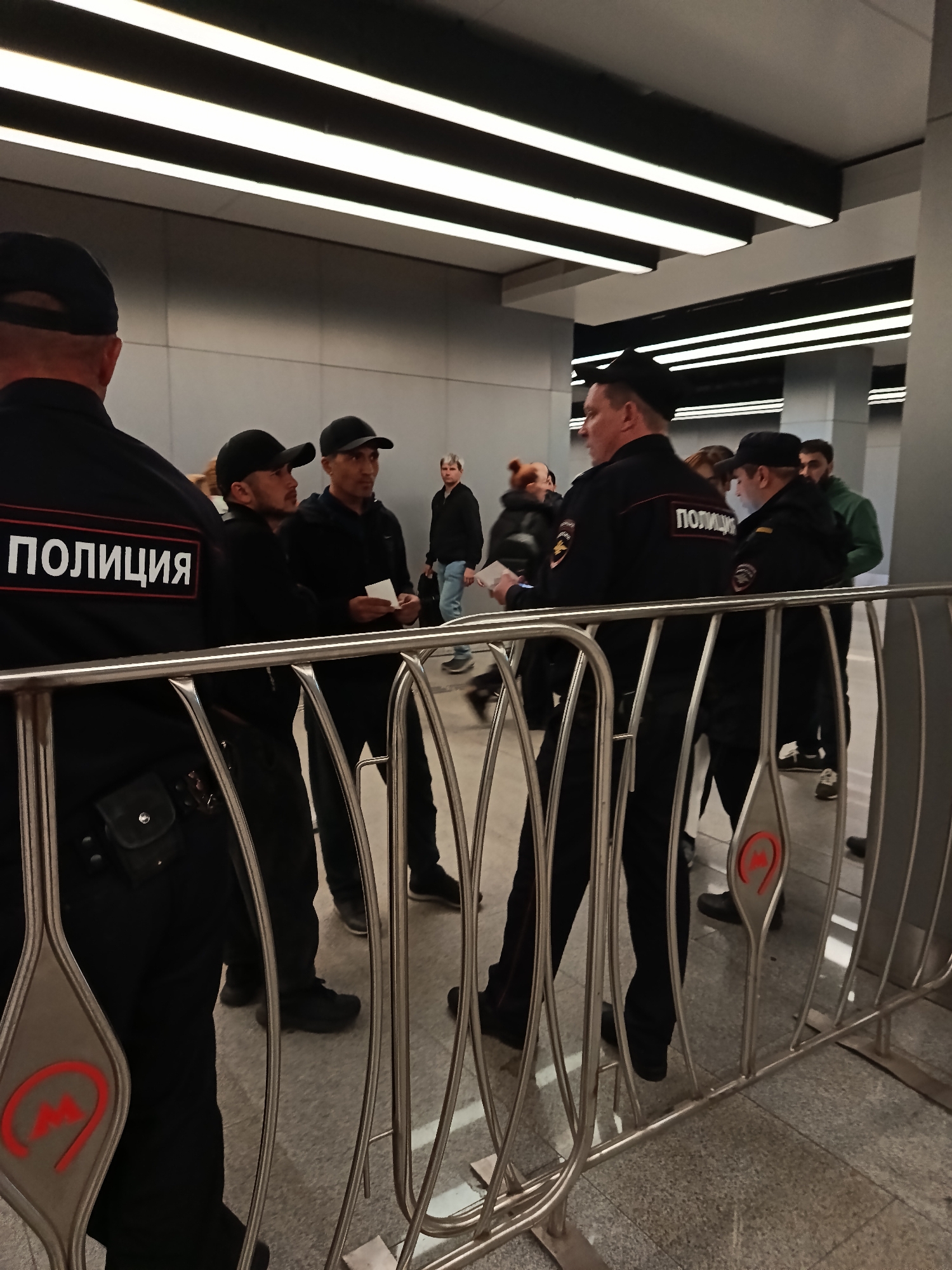 A group of people is standing behind a metal barrier with red logos resembling an "M" inside a teardrop shape, indicating this is the entrance to a metro station in an unknown city. Two men in black are holding pieces of paper and appear to be speaking with a uniformed officer who is holding a similar paper. The officers have the word "ПОЛИЦИЯ" on their backs, identifying them as police. Other people are walking in the background, suggesting a busy environment. The lighting is artificial and bright, coming from a linear light fixture above, illuminating a modern-looking entryway to the metro system.