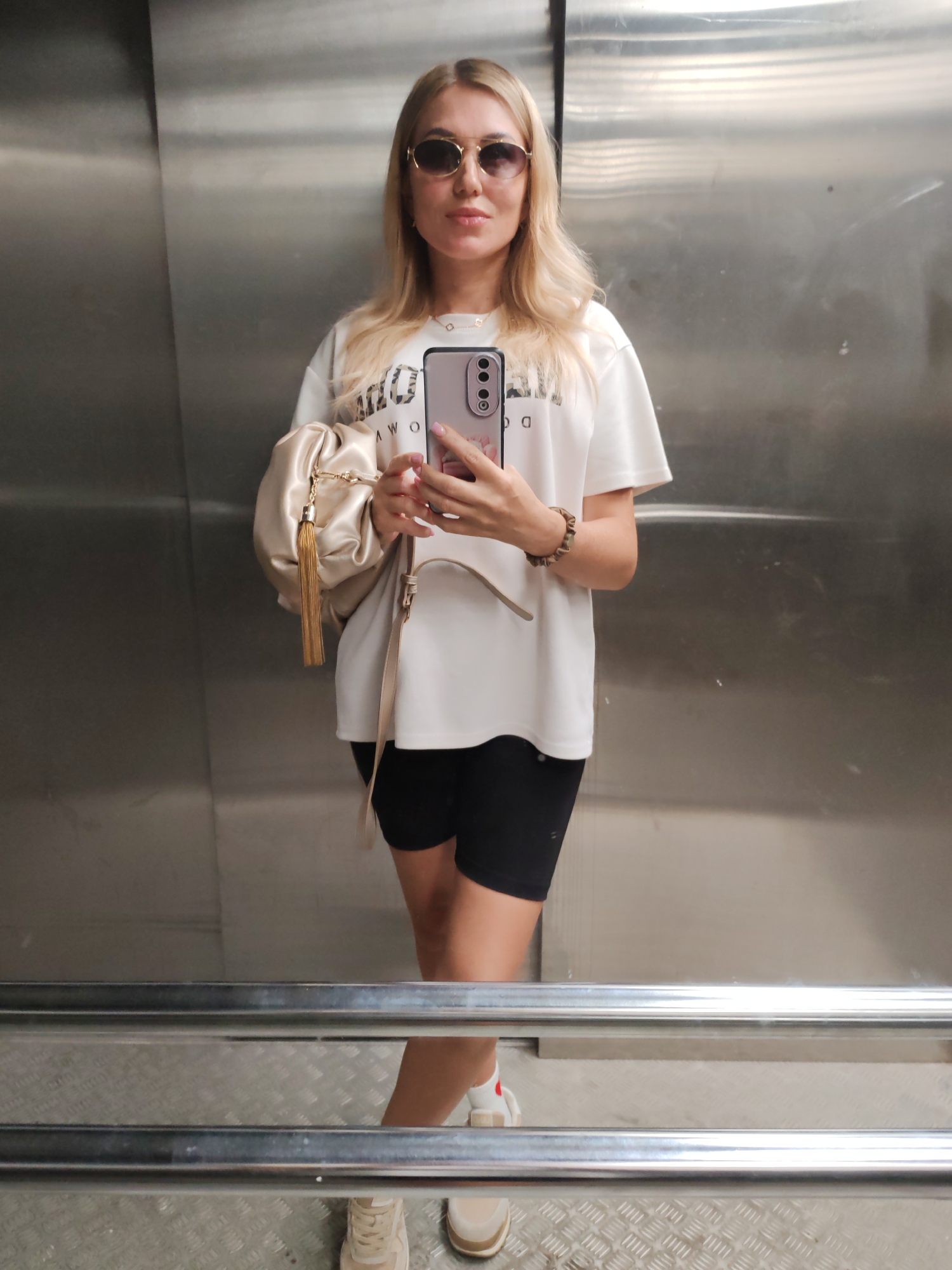 A young woman with blonde hair and sunglasses is taking a mirror selfie in an elevator. She is wearing a white t-shirt, black biker shorts, and beige sneakers. She is holding a beige clutch bag with a gold tassel in her left hand and her phone in her right hand. The elevator walls are made of brushed metal, and the floor is a metal diamond plate pattern. The overall impression is one of a casual, everyday setting. The specific location of Komsomolsk-on-Amur, Russia, is not discernible from the visual elements of the image.