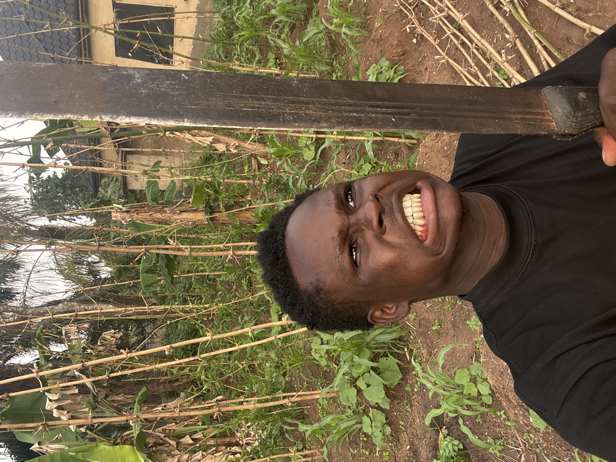 A man is holding a machete in an outdoor scene, likely in the city of Aboh, Nigeria. The man is positioned in the foreground, and vegetation is visible around him, along with some bamboo-like structures. A building is also discernible in the background. The man is smiling or grinning, and the machete is held in a way that it partly obscures the background. The ground appears to be dirt or soil.