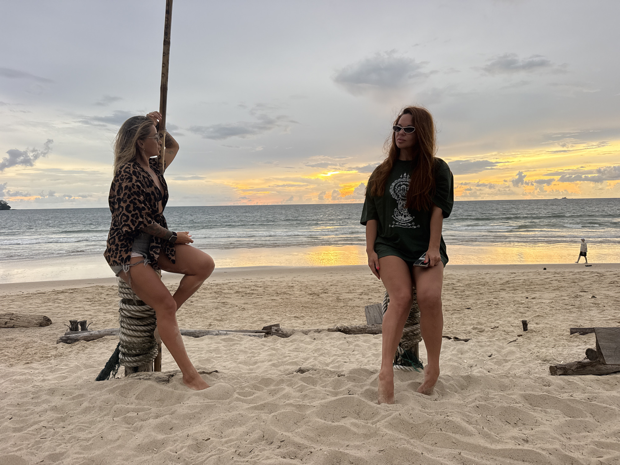 Two women are sitting on a sandy beach in Choeng Thale, Thailand. The woman on the left is sitting on a thick rope-wrapped pole, leaning against a wooden post. She has blonde hair and is wearing sunglasses, a leopard-print kimono, and denim shorts. The woman on the right is sitting on a similar rope-wrapped pole, facing the ocean. She has long brown hair and is wearing white rectangular sunglasses and a dark green t-shirt with a graphic print.
The scene is set during sunset, with a colorful sky casting a warm glow over the ocean. The waves are gently lapping at the shore, and a single person is walking in the distance. The beach is sandy with some driftwood scattered around. The overall mood is relaxed and serene, suggesting a peaceful evening by the sea. The graphic on the woman's t-shirt is a circular design with intricate details.