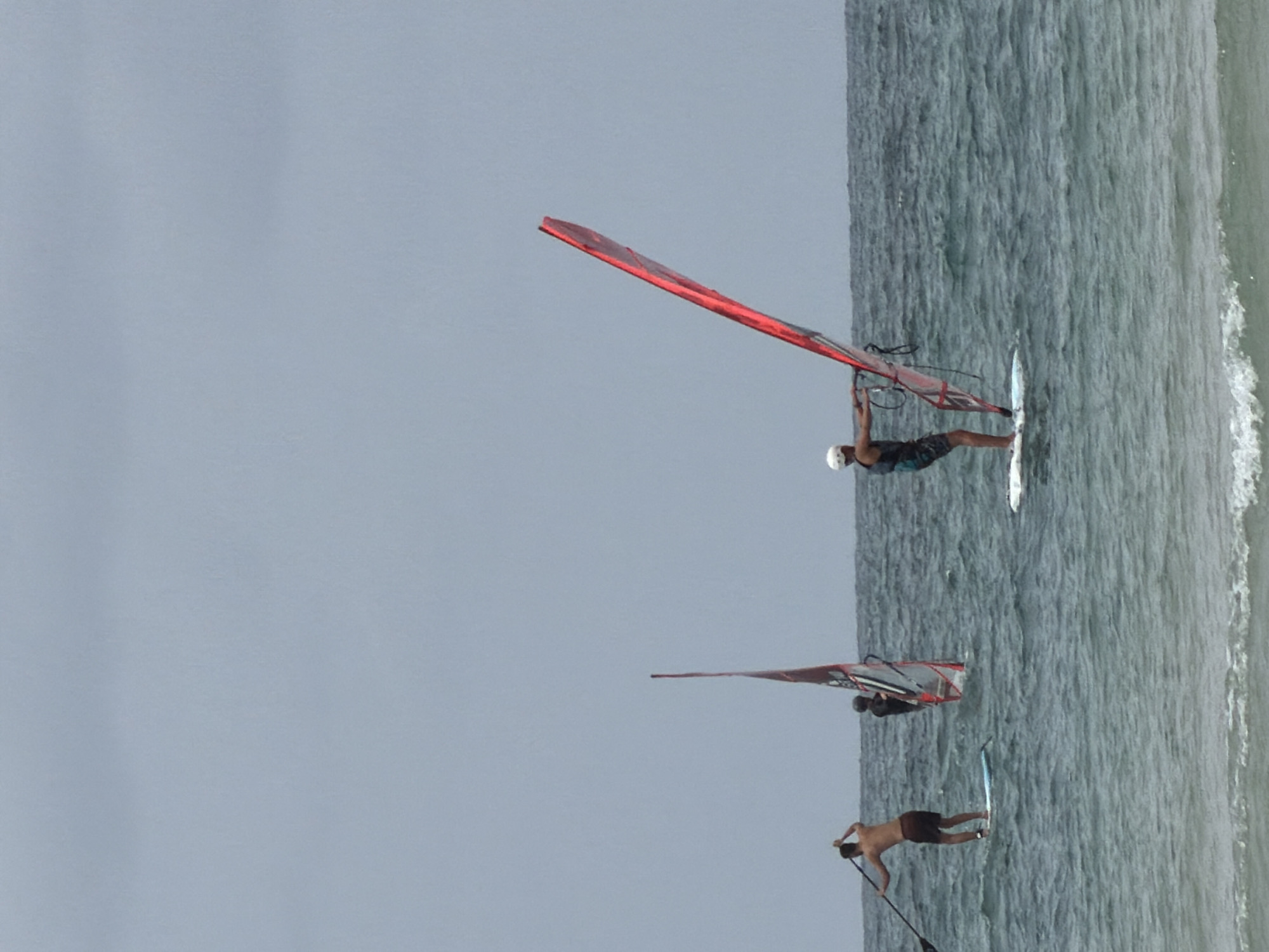 The image depicts three individuals engaged in water sports off the coast of Tel Aviv, Israel. The first person is windsurfing, utilizing a red sail. The second person is also windsurfing and is farther away. The third person appears to be paddleboarding. The sky is overcast, and the water is a mix of dark and light shades, suggesting varying depths or wave activity. The shoreline is visible on the right.