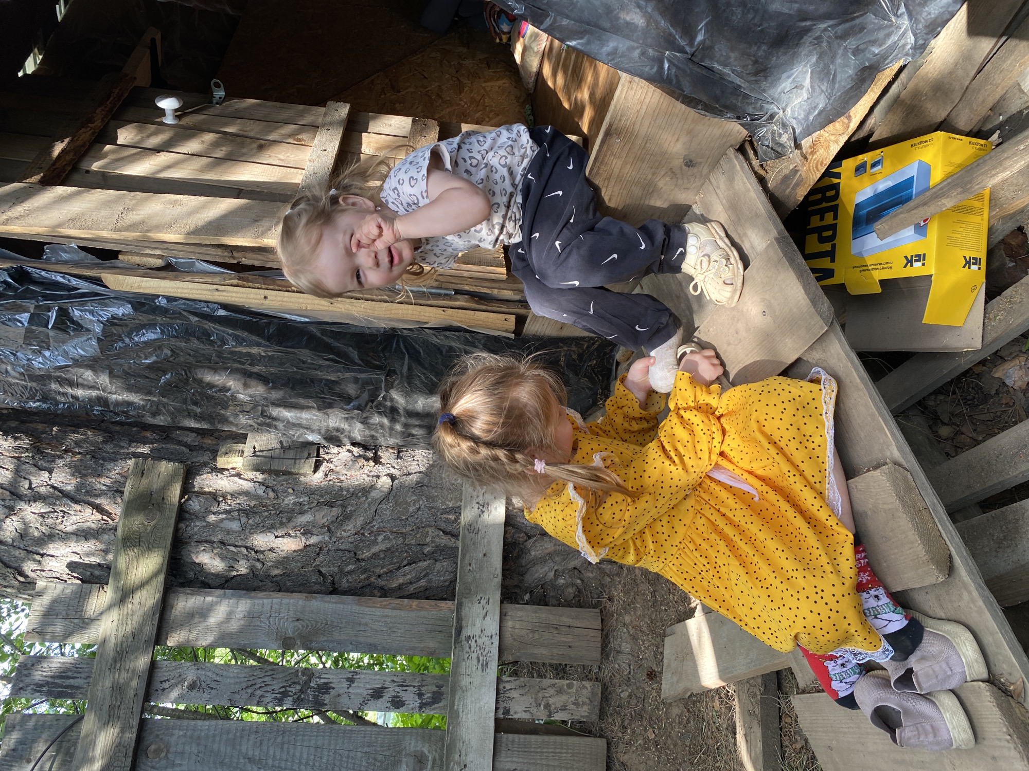 The image depicts two young girls in an outdoor setting, likely in Chaikovsky, Russia. The older girl, dressed in a yellow polka dot dress and wearing socks with a Christmas theme and grey tennis shoes, sits on a wooden structure. The younger girl is lying on her back on a similar wooden structure. She is wearing a white shirt with light grey spots, black pants with white logos, and light-colored tennis shoes.
The scene includes various objects such as wooden planks, a yellow electrical box, and a dark plastic tarp. The environment is sunny. The girls appear to be interacting in a playful manner, with the younger girl holding her hand near her face.