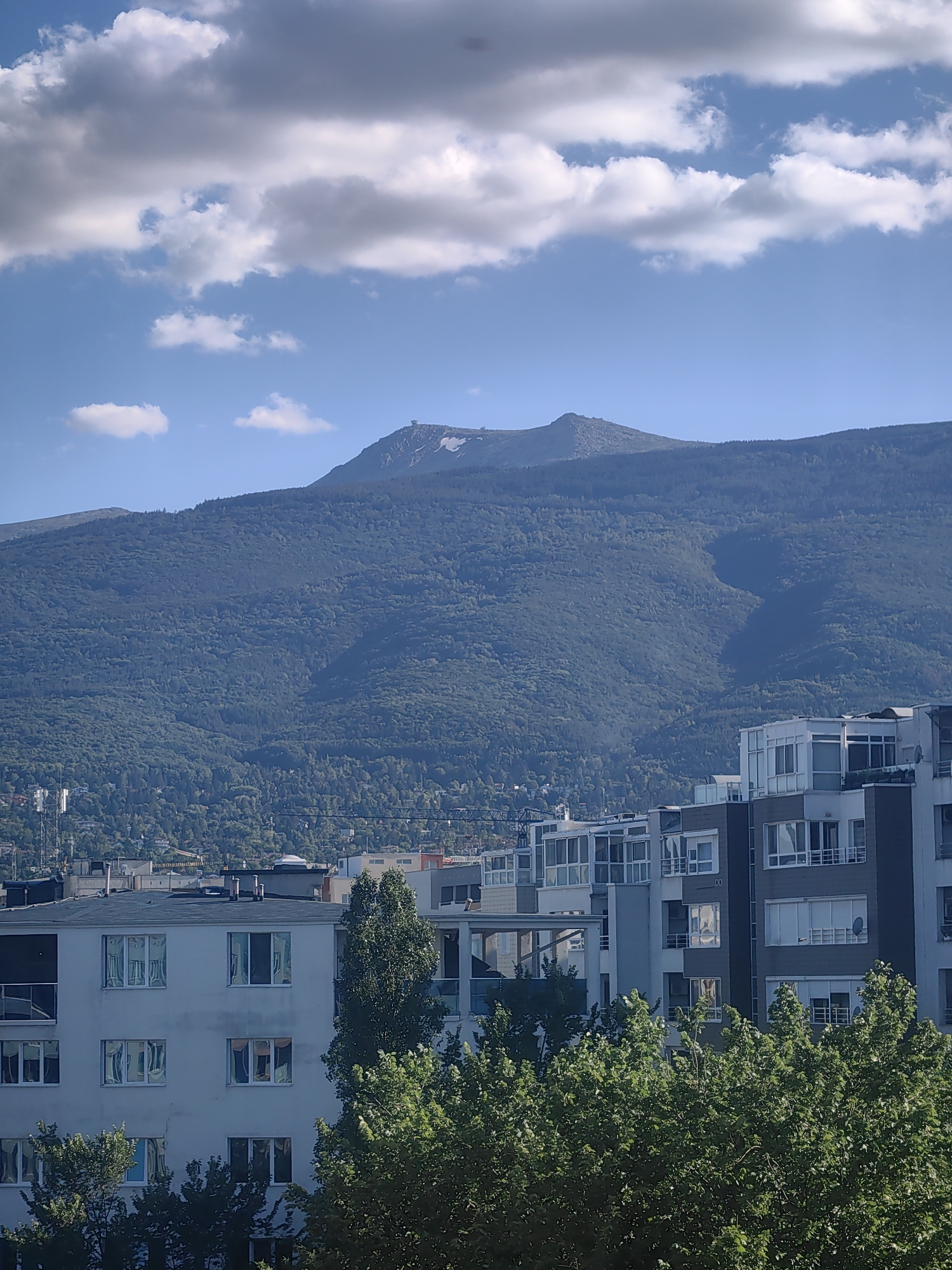 The image captures a scenic view of Vitosha Mountain overlooking the city of Sofia, Bulgaria. The foreground features several apartment buildings with modern architecture, characterized by balconies and large windows. Lush green trees obscure parts of the lower floors of these buildings. In the midground, the dense, forested slopes of Vitosha Mountain rise majestically, with a visible shadow cast across its surface. Near the summit of the mountain, a structure, likely an observatory or a tower, is discernible. The sky above is a clear blue, punctuated by scattered white clouds, some of which are large and fluffy, while others are more wispy. The overall impression is one of urban development encroaching upon a natural landscape under a bright, partly cloudy sky.