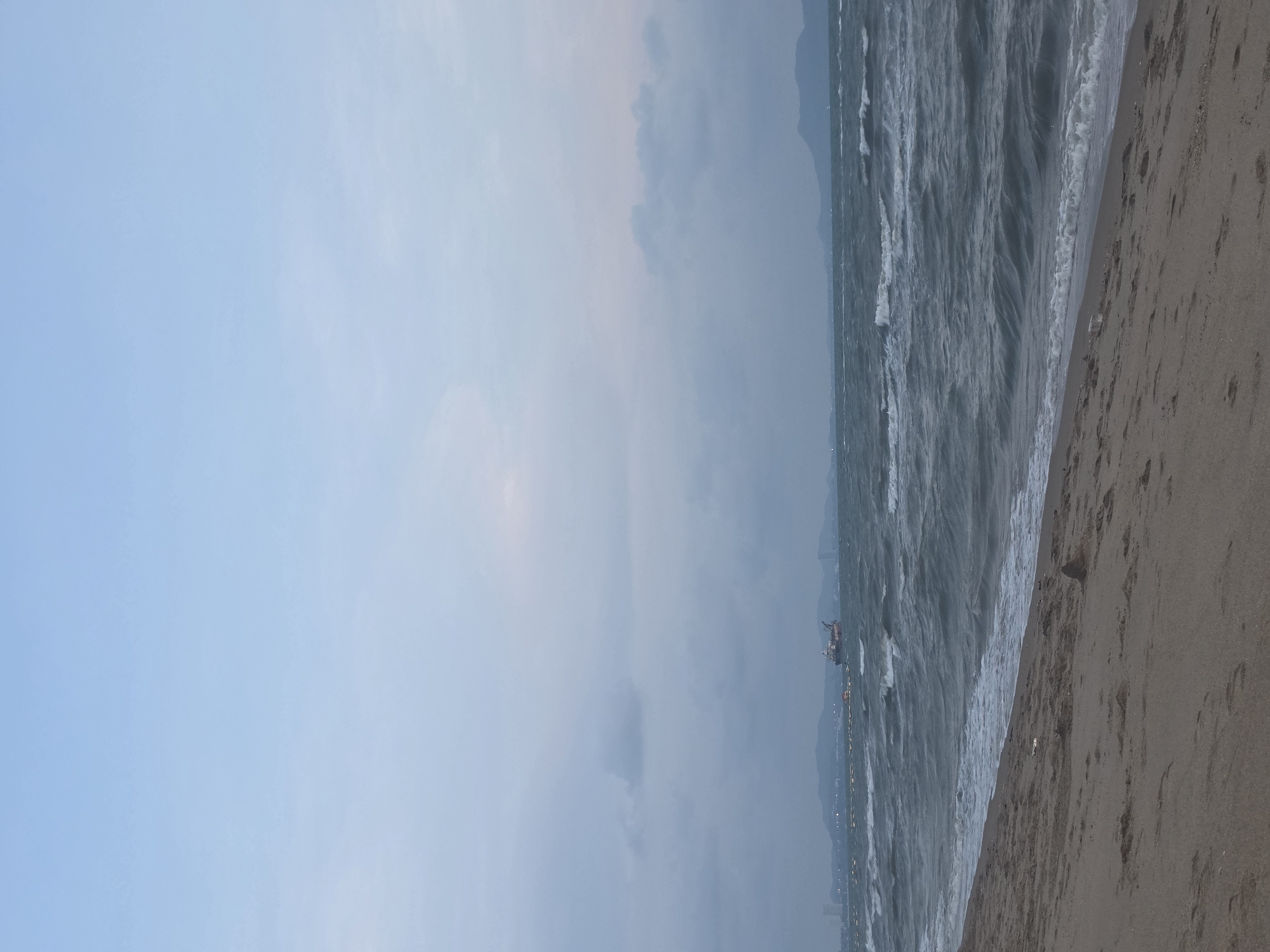 The image displays a wide coastal landscape, oriented vertically, depicting a serene yet active beachfront scene at what appears to be dawn or dusk.
The left portion of the image is dominated by the sky, which transitions from a soft, pale blue at the top to a more muted, hazy grey-blue closer to the horizon, suggesting an overcast or diffused light condition. The central section features the expansive ocean, characterized by choppy, dark greyish-blue water with numerous white-capped waves rolling towards the shore.
In the mid-distance on the water, a prominent dark vessel, possibly a cargo ship or an offshore platform, can be seen. Along the distant horizon, a faint landmass of mountains or hills is visible, partially obscured by the atmospheric haze. A string of small, bright lights dots the distant coastline, indicating either coastal city lights or illuminated structures like piers or breakwaters.
The right side of the image reveals a sandy beach of a light brown color, where the waves gently break and recede. Faint indentations, possibly footprints, are discernible on the sand. No specific human activity is in progress on the immediate beach, but the distant lights and vessels imply human presence along the coast.
The overall mood is tranquil yet dynamic, highlighting the natural interaction between the sea and the shore under a soft, diffused light. There is no visible text in the image. The setting is consistent with a coastal location like Pattaya City, Thailand, known for its beaches and maritime activity.