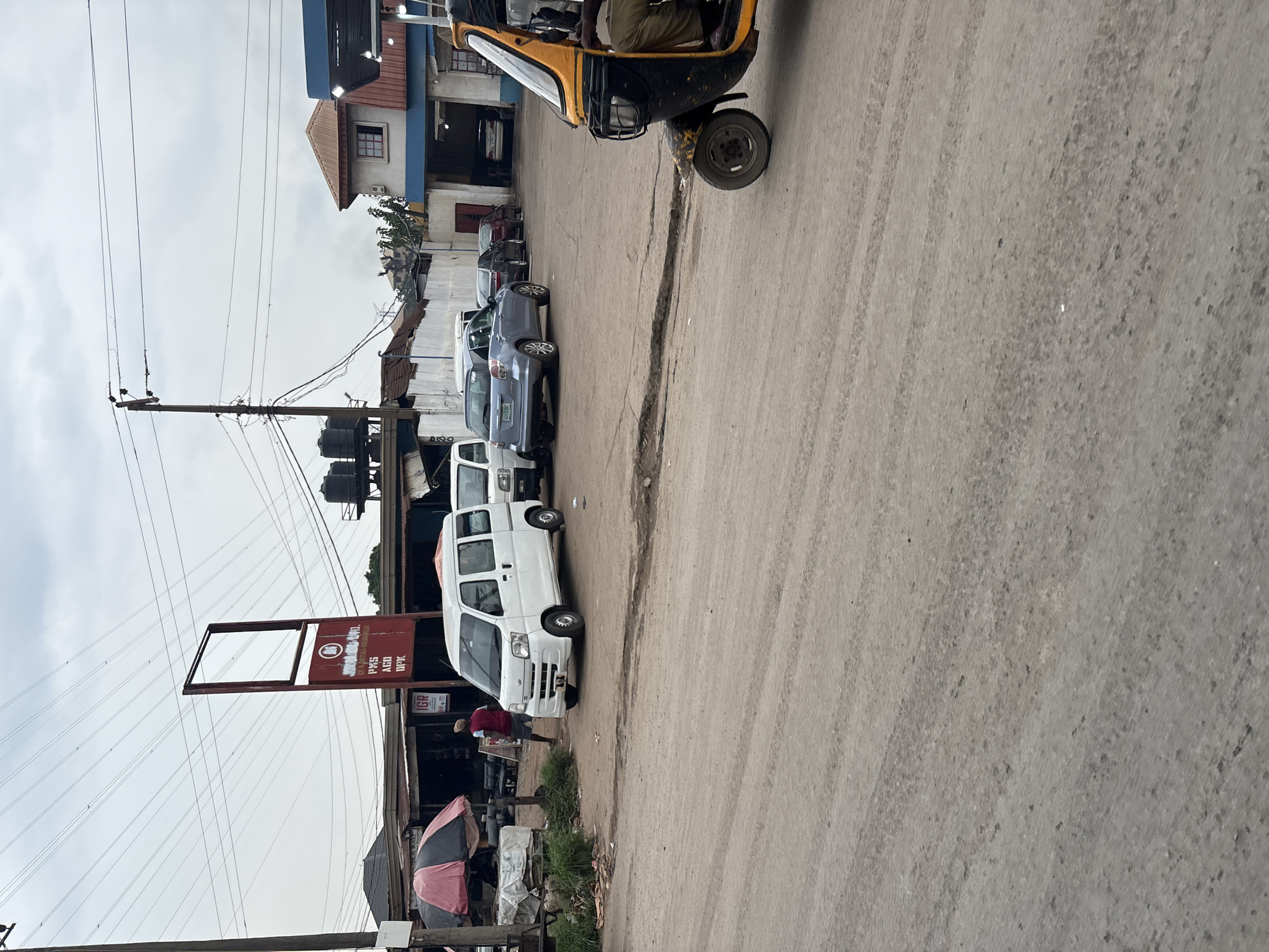 The image captures a street scene in Onitsha, Nigeria, likely near a commercial area. In the foreground, a dirt road displays tire tracks and scattered debris. To the right, a yellow and black auto-rickshaw, also known as a Keke, is partially visible, with a driver seated inside wearing khaki trousers. Further into the scene, several vehicles are parked or driving on the road. A white minivan is prominent, with a man in a red shirt and blue shorts standing near it. Behind the minivan, other cars, including a blue hatchback and a dark-colored sedan, are visible. Several market stalls or shops with various goods are set up along the side of the road, some covered by umbrellas. A tall, red signpost stands empty at the top, with text and a circular logo on its lower section. Overhead, a network of power lines crisscrosses the cloudy sky, supported by utility poles. Buildings, including a multi-story structure with a reddish roof and a workshop with an open bay, are present in the background. Water tanks are also visible on top of some structures. The overall impression is a bustling, everyday street life in a Nigerian city.