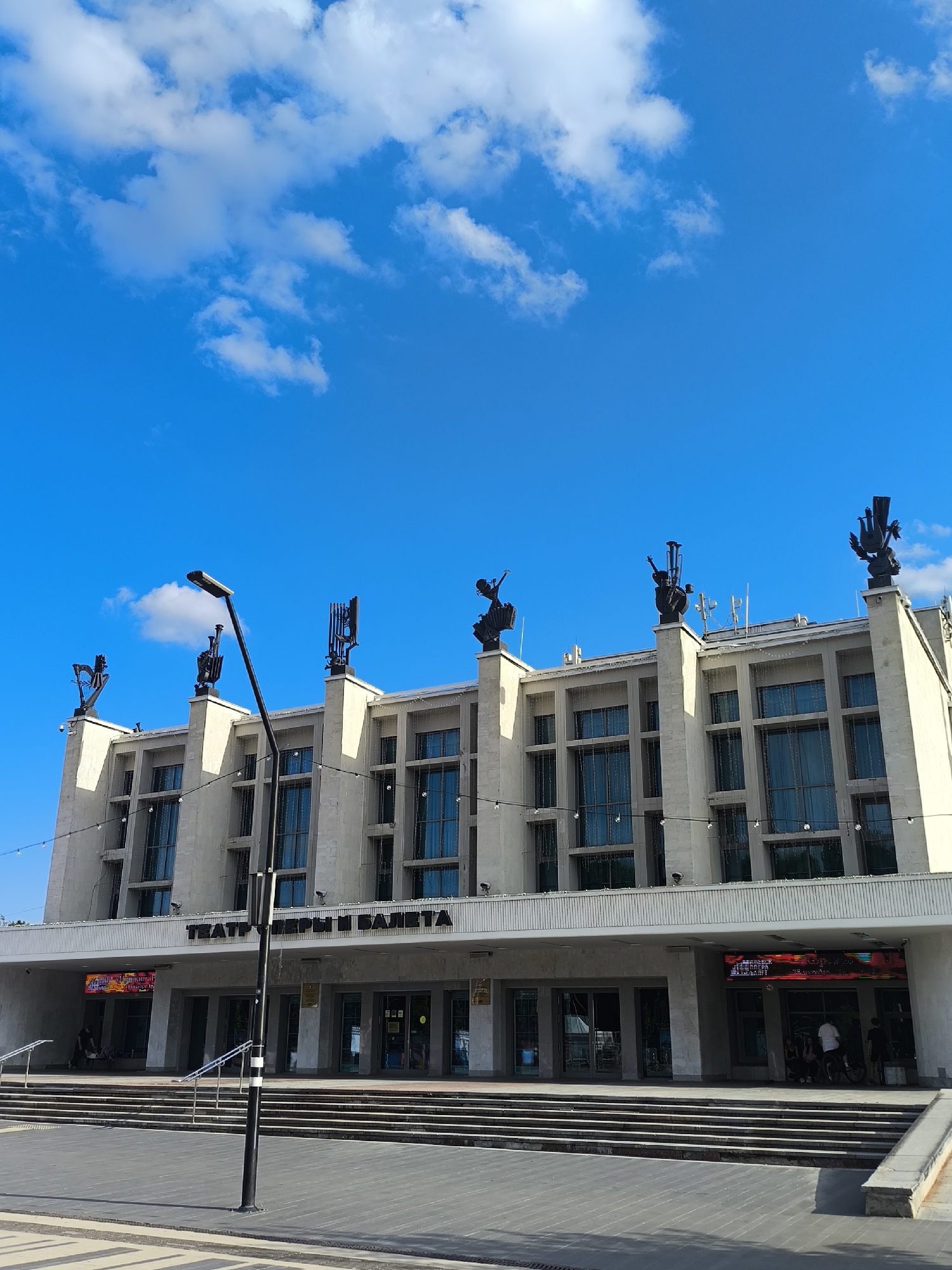 The image shows the State Opera and Ballet Theatre in Izhevsk, Russia, on a bright, sunny day with a blue sky dotted with white clouds. The grand, modern building features a light-colored facade with tall, narrow windows. Decorative sculptures, some resembling musical instruments, are perched atop pillars on the roofline. A prominent street lamp stands in the foreground, and a wide set of stairs leads up to the theatre's entrance. Several entrances with glass doors are visible, and a digital display board above one of them shows colorful graphics. A few people are gathered near the entrance, and a bicycle is parked nearby. The overall scene is one of civic grandeur and public space.
