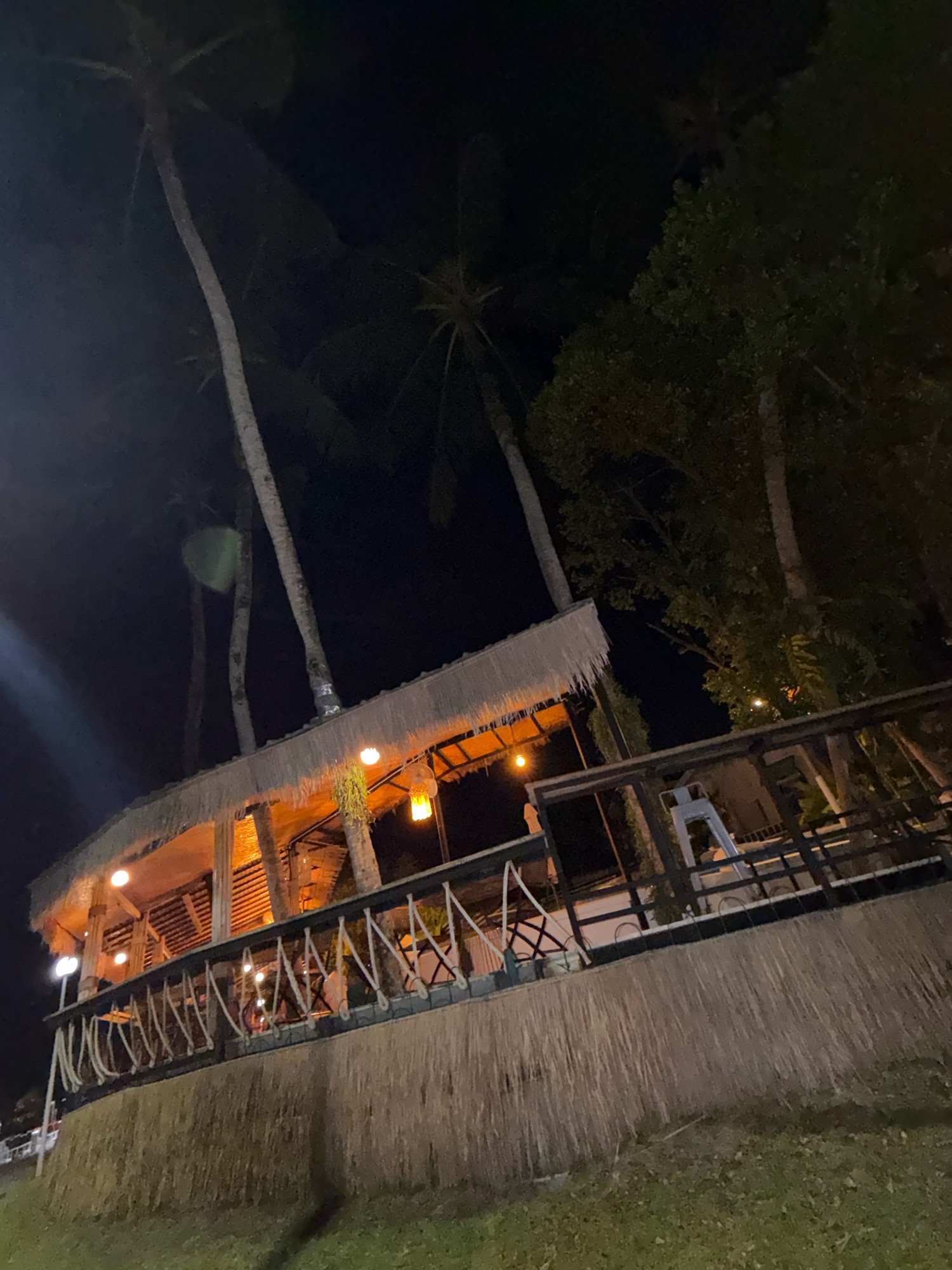 This is a nighttime exterior, low-angle shot of a thatched-roof structure elevated on stilts. The structure, which appears to be a restaurant or bar, is illuminated from within by warm, yellow lights. The roof is made of dried palm leaves, and the railings of the elevated deck are made of wood and rope. Palm trees are visible behind the structure, and a dense canopy of dark green trees is on the right side of the frame. The ground in front of the structure is covered with grass. The scene is set in Ko Kaeo, Thailand, and evokes a tropical, evening atmosphere. There are no people visible in the image.