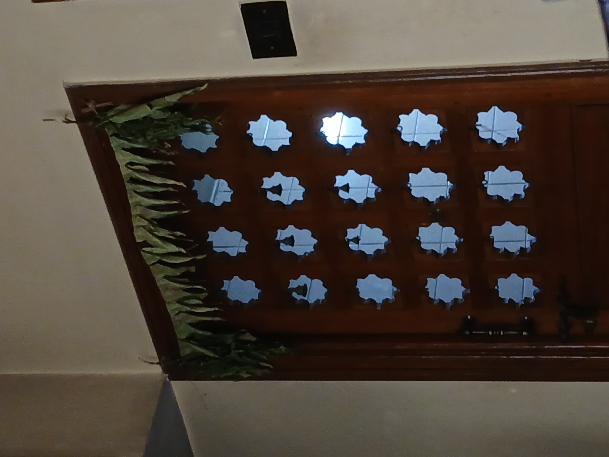 A low-angle, indoor shot depicts a wooden window with decorative floral cutouts, embellished with greenery on its left side. Above the window, partially visible against a light-colored wall, is a dark, rectangular object, possibly a switch or vent. The window's floral cutouts reveal glimpses of the exterior. Below the window, a light-colored wall extends into the frame, suggesting an interior space. The location is inferred to be Bengaluru, India. The image is static, devoid of people, focusing on architectural and decorative elements.