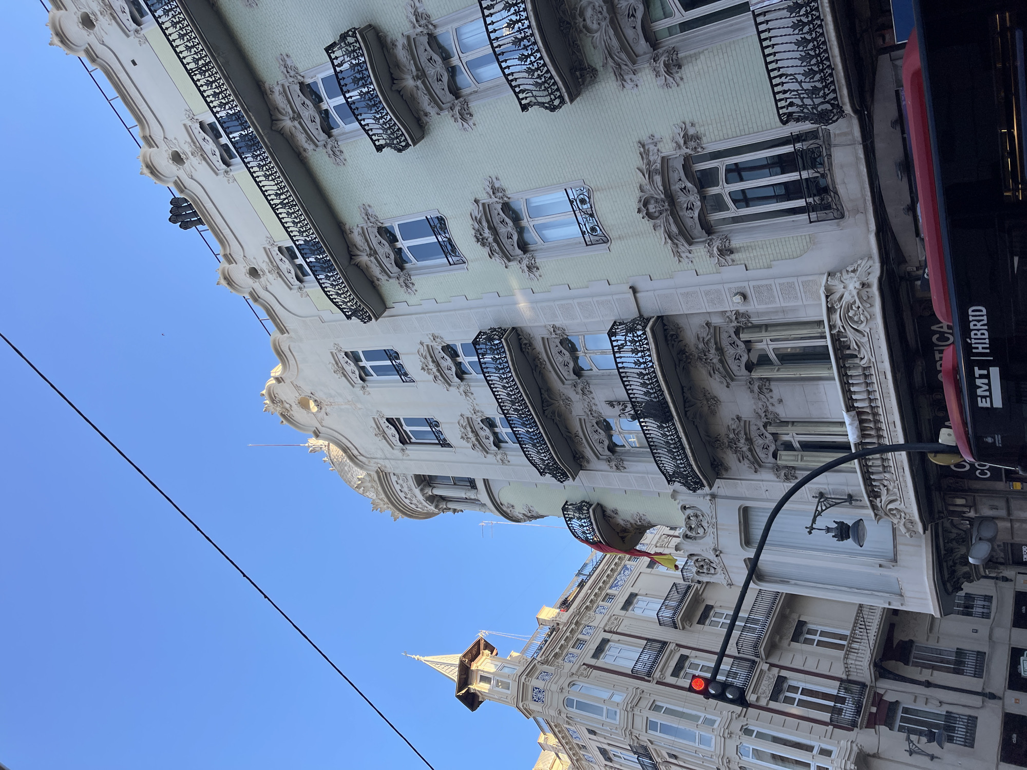 The image shows a vertical view of buildings in València, Spain, with a focus on architectural details. The sky is clear and blue. The buildings display ornate designs, including intricate window frames, balconies with decorative iron railings, and carved facades. One building has a light green hue. A traffic light is visible, indicating a street. Part of a bus with the word "HÍBRID" is also visible. The architectural style is reminiscent of early 20th-century European design, with elements of Art Nouveau. The buildings' orientations suggest they are facing a relatively narrow street.