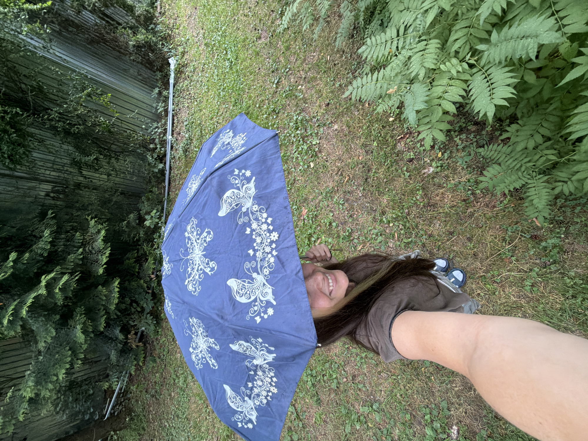 A high-angle, full-body selfie shows a smiling young woman holding a dark blue umbrella with white butterfly and floral patterns above her head. She is outdoors on a grassy area with some sparse weeds and fallen pinecones. To the right, a large patch of green ferns is visible. In the background, a wooden fence and a dense cluster of evergreen trees form the backdrop. The woman is wearing a brown t-shirt and grey shorts. Her left arm, extended to take the photo, is prominent in the foreground. The image appears to be taken in Udelnaya, Russia, given the presence of evergreen trees typical of the region.