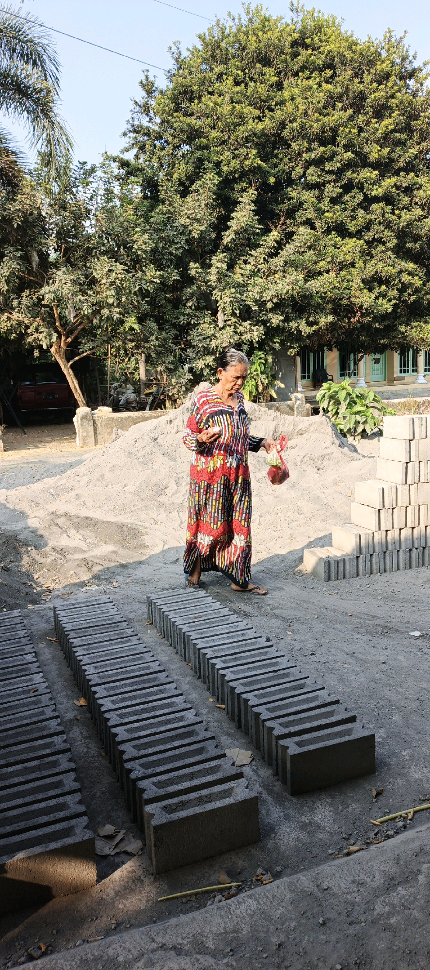 An elderly woman with gray hair pulled back walks across a construction site in Kasiyan, Indonesia. She is wearing a vibrant, long-sleeved, patterned dress in red, yellow, white, and black. In her right hand, she carries a clear plastic bag containing what appears to be red fruit, and in her left hand, she holds a small white object. The ground is a mixture of concrete and sand, with rows of gray concrete blocks neatly stacked in the foreground and to the right. Behind her, a large pile of sand dominates the mid-ground. Lush green trees, including palm trees, provide a natural backdrop. In the far background, a red vehicle is parked near a building with a green roof and white pillars. The scene is bathed in the warm light of what appears to be late afternoon sun.