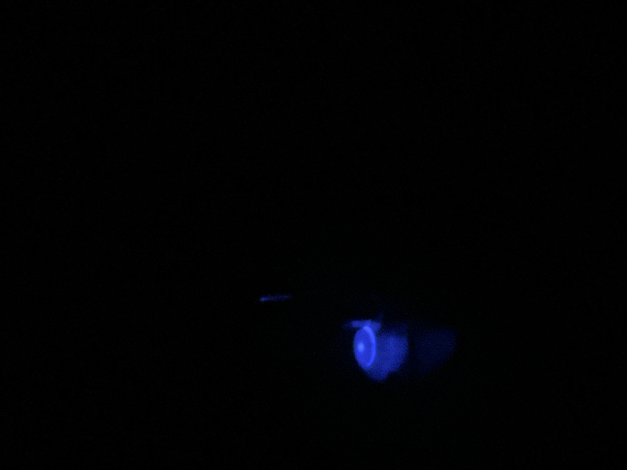 The image shows a dark scene with a single, small, bright blue light source in the lower left quadrant. The light appears to emanate from a small object, possibly a button or a small LED, and has a slightly diffused halo around it. There is a faint, vertical streak of light above and to the right of the primary light source, suggesting another, less intense light or a reflection. The rest of the image is completely black, indicating a very dark environment. No specific location in Krakow, Poland, can be identified from the visual information provided. The scene is devoid of any human presence, activity, or discernible environmental features beyond the darkness and the light sources.