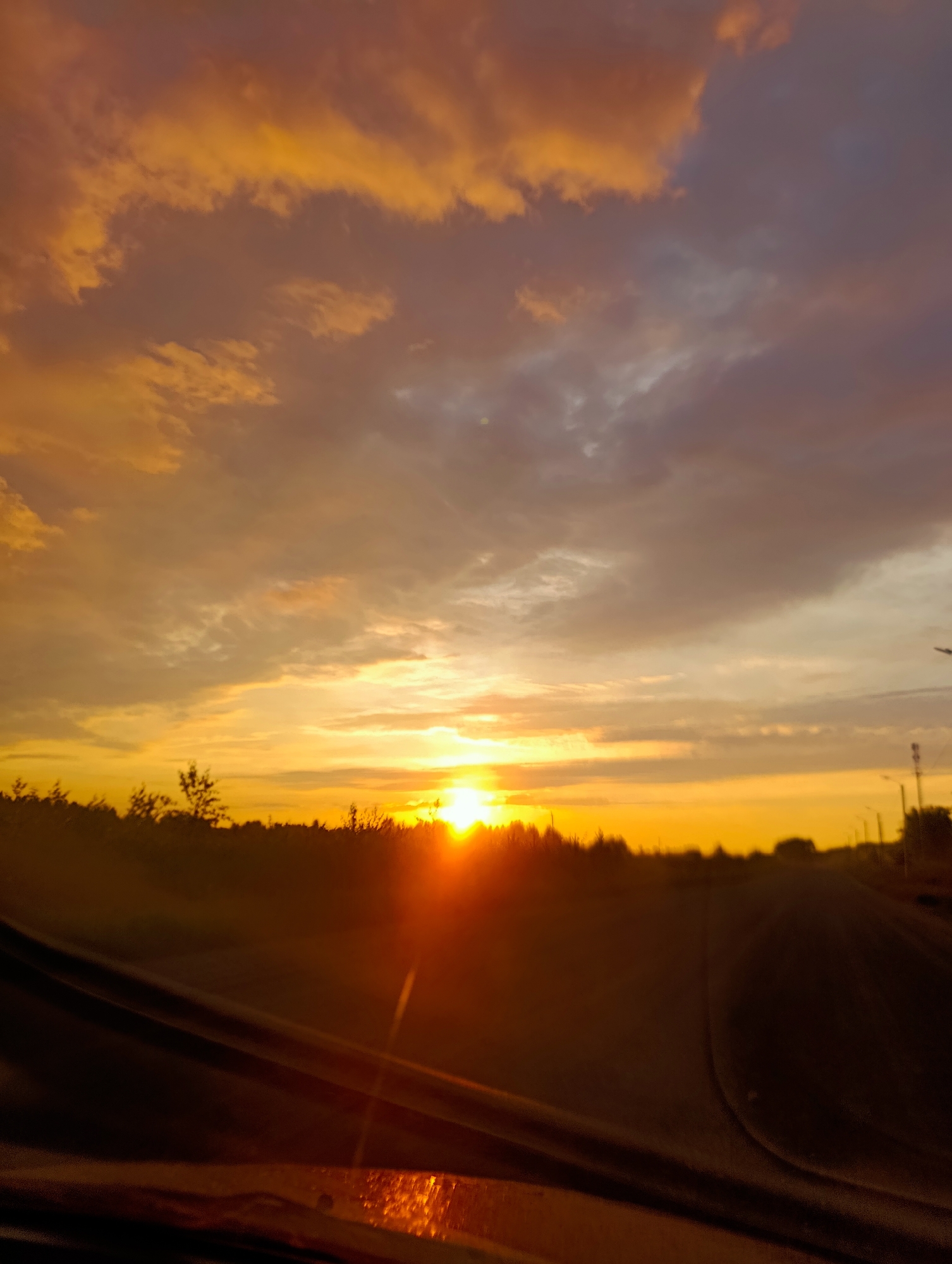 A car dashboard is visible in the foreground, with sunlight reflecting off its surface. Beyond the car, a road stretches into the distance, bordered by trees on the left and illuminated by the setting sun. The sky is filled with dramatic clouds in shades of orange, yellow, and purple. The scene is set in the city of Kamennyi Kliuch, Russia.