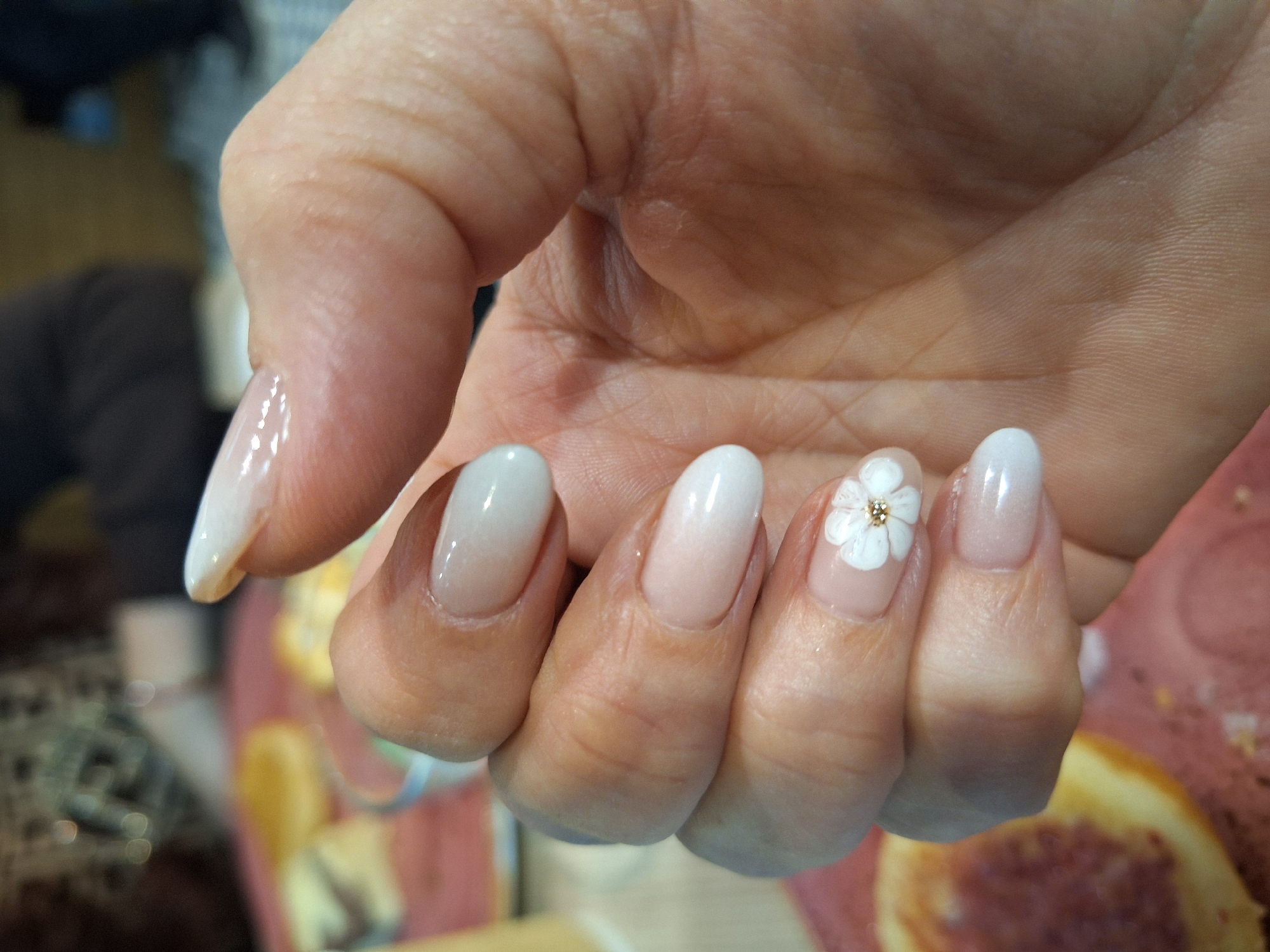 The image shows a close-up of a person's hand with manicured nails. The nails are oval-shaped and appear to have a gradient or ombre effect, transitioning from a nude or light pink base to a white tip. The ring finger has a small, white flower design with a gold-colored center. The hand is held against a blurred background, partially showing a table or surface with food items or decorative objects. There is no clear evidence to determine if it's in Wrocław, Poland.
