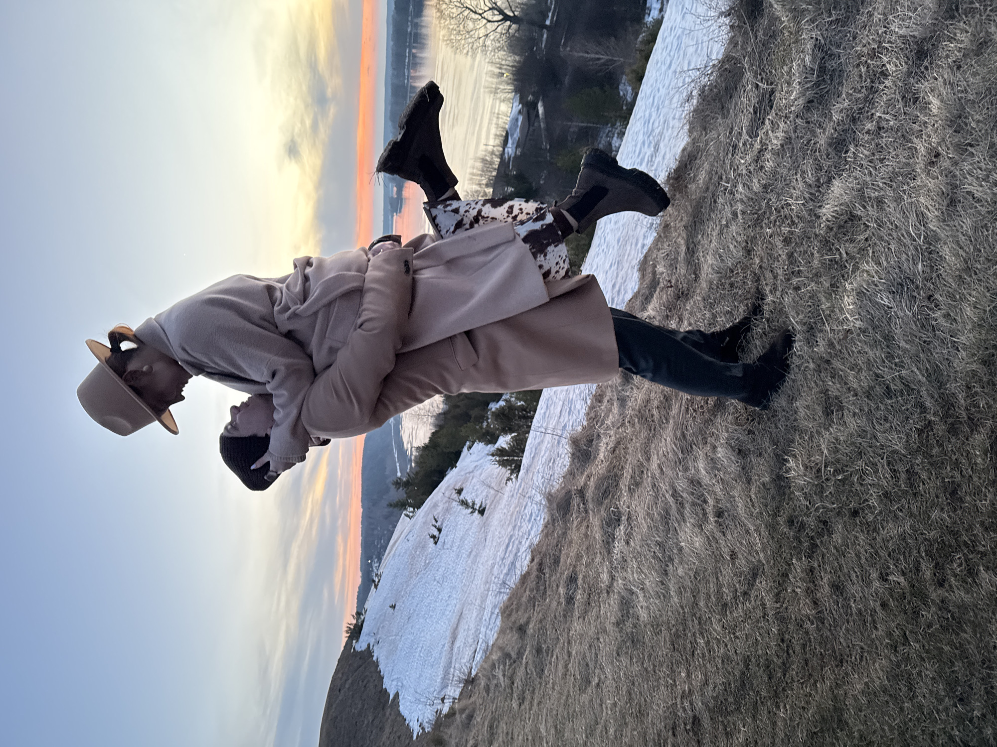 A photograph shows two people embracing on a snow and grass-covered hillside in Ольховка, Russia, at sunset. The person on the left wears a tan hat and a tan coat. The person on the right wears a black beanie and a beige coat. The location overlooks a river, and snow is visible on the hillside and surrounding areas. The sky is transitioning from blue to orange as the sun sets.