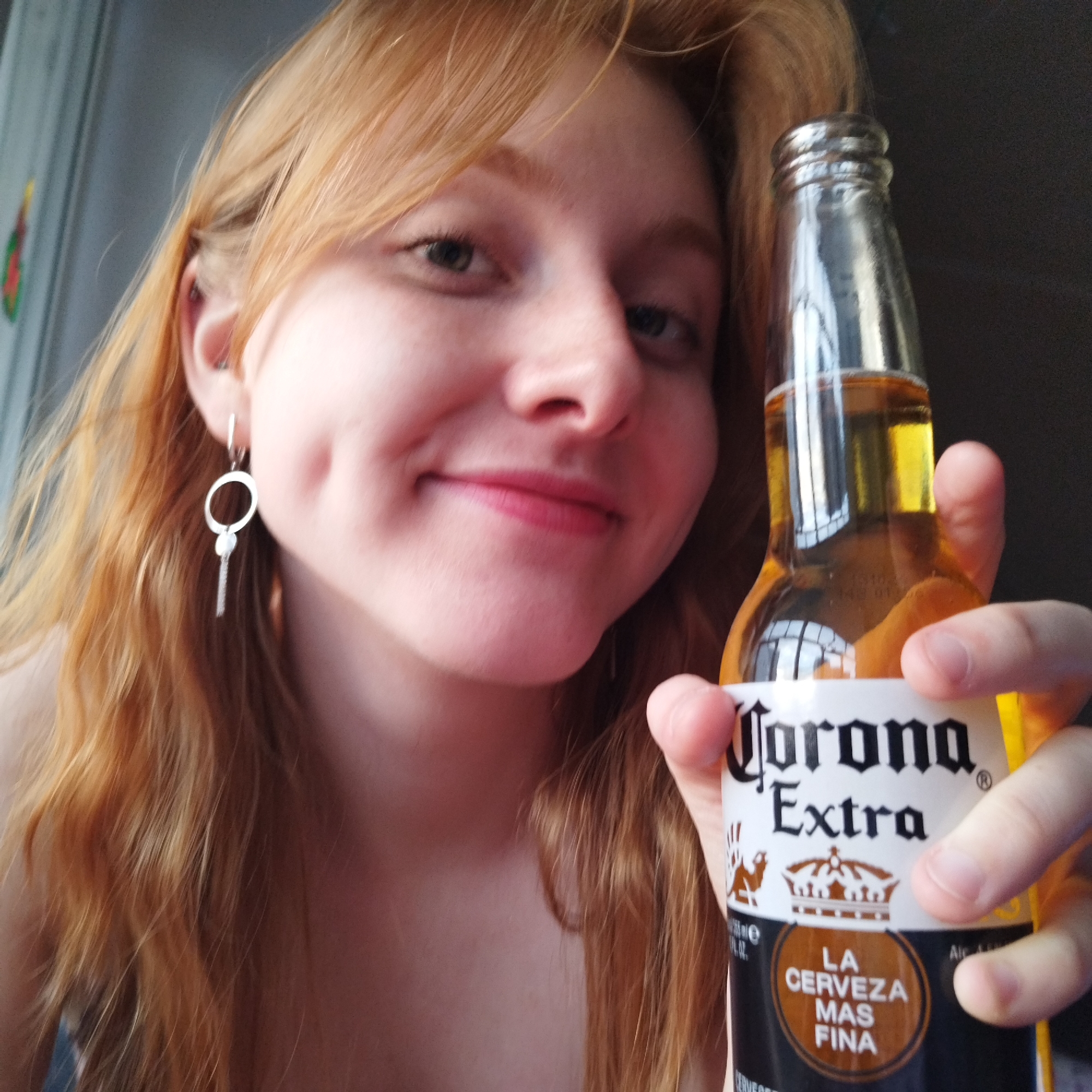 A young woman with long, wavy ginger or strawberry blonde hair is captured in a close-up, chest-up selfie. She is smiling or smirking directly at the camera, revealing prominent dimples on her cheeks and a hint of pink lipstick. Her left ear is visible, adorned with a silver-colored earring consisting of a small ring and a dangling bar. Her eyes appear to be light-colored.
She holds a clear glass bottle of Corona Extra beer in her right hand, positioned prominently in the foreground. The bottle contains a golden-colored liquid and is approximately one-third full. The white label on the bottle clearly displays the brand "Corona Extra" in black gothic script. Below the brand name, a brown circular emblem features the text "LA CERVEZA MAS FINA" (The Finest Beer). Other visible text on the label includes "355 ml e", "12 fl. oz.", "Alc Vol", and the beginning of "CERVEZA". The characteristic Corona crown logo and a small camel logo are also discernible on the label.
The background is softly blurred, appearing in shades of grey and dark blue, suggesting an indoor setting. A faint, blurry splash of red and green is visible on the far left, possibly indicating a decoration. The overall impression is one of casual cheerfulness. This scene takes place in Yekaterinburg, Russia.
