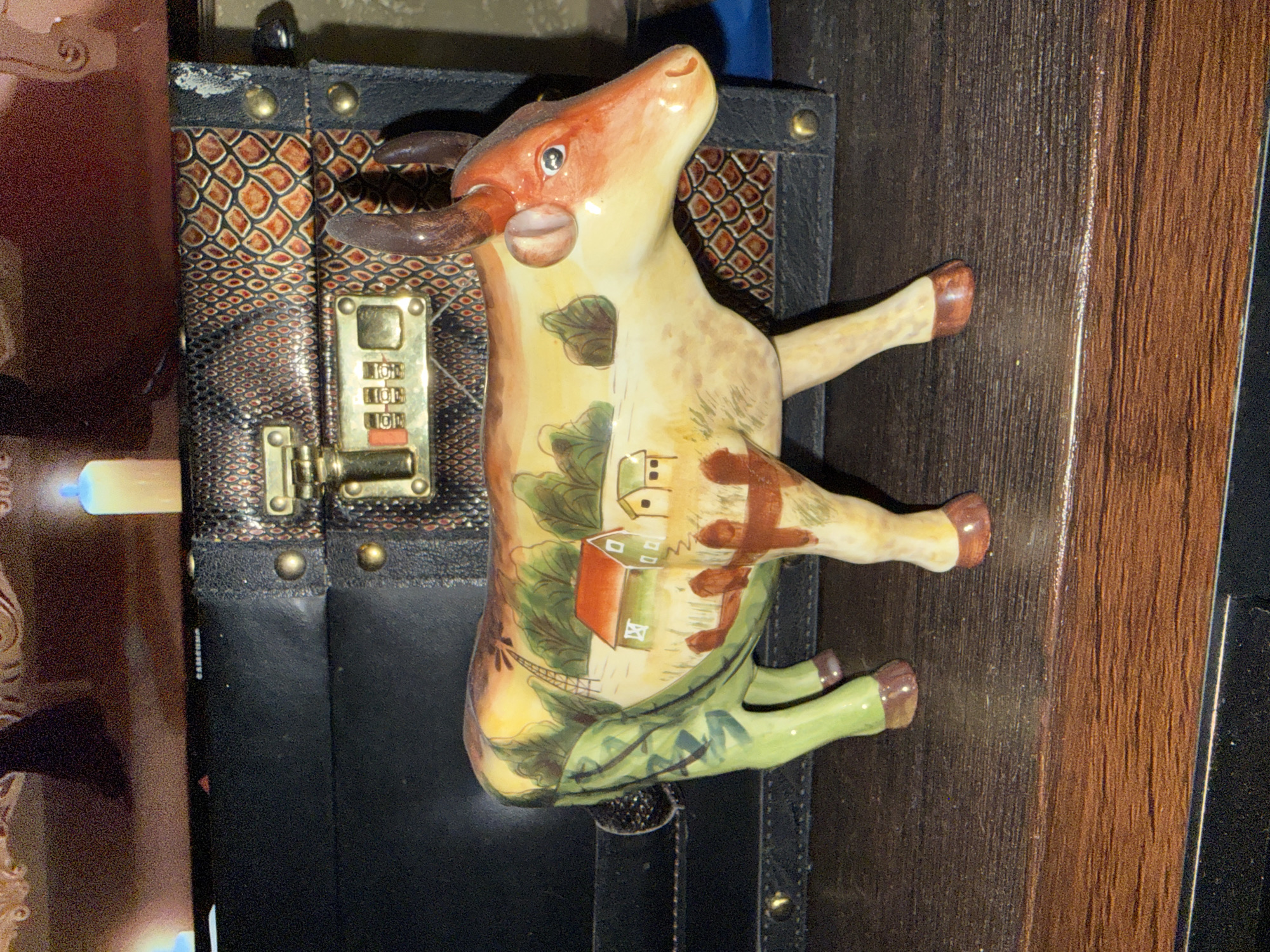 A ceramic cow figurine with a pastoral landscape painted on its body stands on a wooden surface in front of a black and textured case with a combination lock. The figurine depicts rolling hills, houses, trees, and a windmill. The case is adorned with gold-colored hardware. The background shows a blurry image of what appears to be a screen or picture with two lit candles.