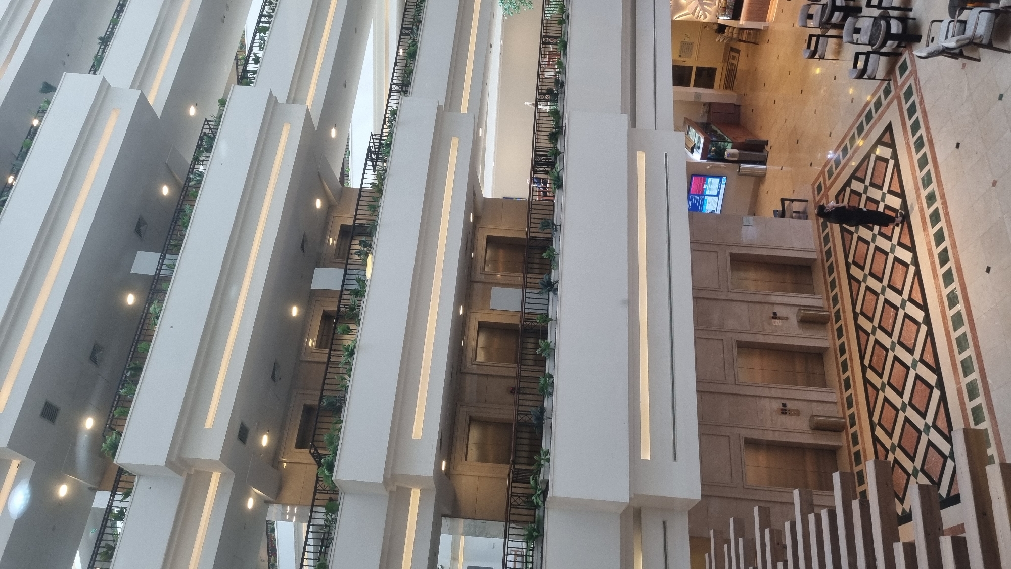 The image depicts the interior of a multi-story atrium, likely a hotel or a large corporate building. The architecture is modern, with clean lines and multiple levels featuring balconies and walkways. Plants are strategically placed along the railings of these balconies, adding a touch of greenery. Several elevators are visible on different floors, with their metallic doors reflecting the ambient light. The floors are tiled in a geometric pattern, and there are areas with seating arrangements, suggesting a lobby or lounge space. A woman in dark clothing is walking across the patterned floor in the foreground, appearing to be on her phone. The overall impression is one of a spacious and well-lit interior environment. There is no discernible information in the image to place it specifically in Tel-Aviv, Israel.