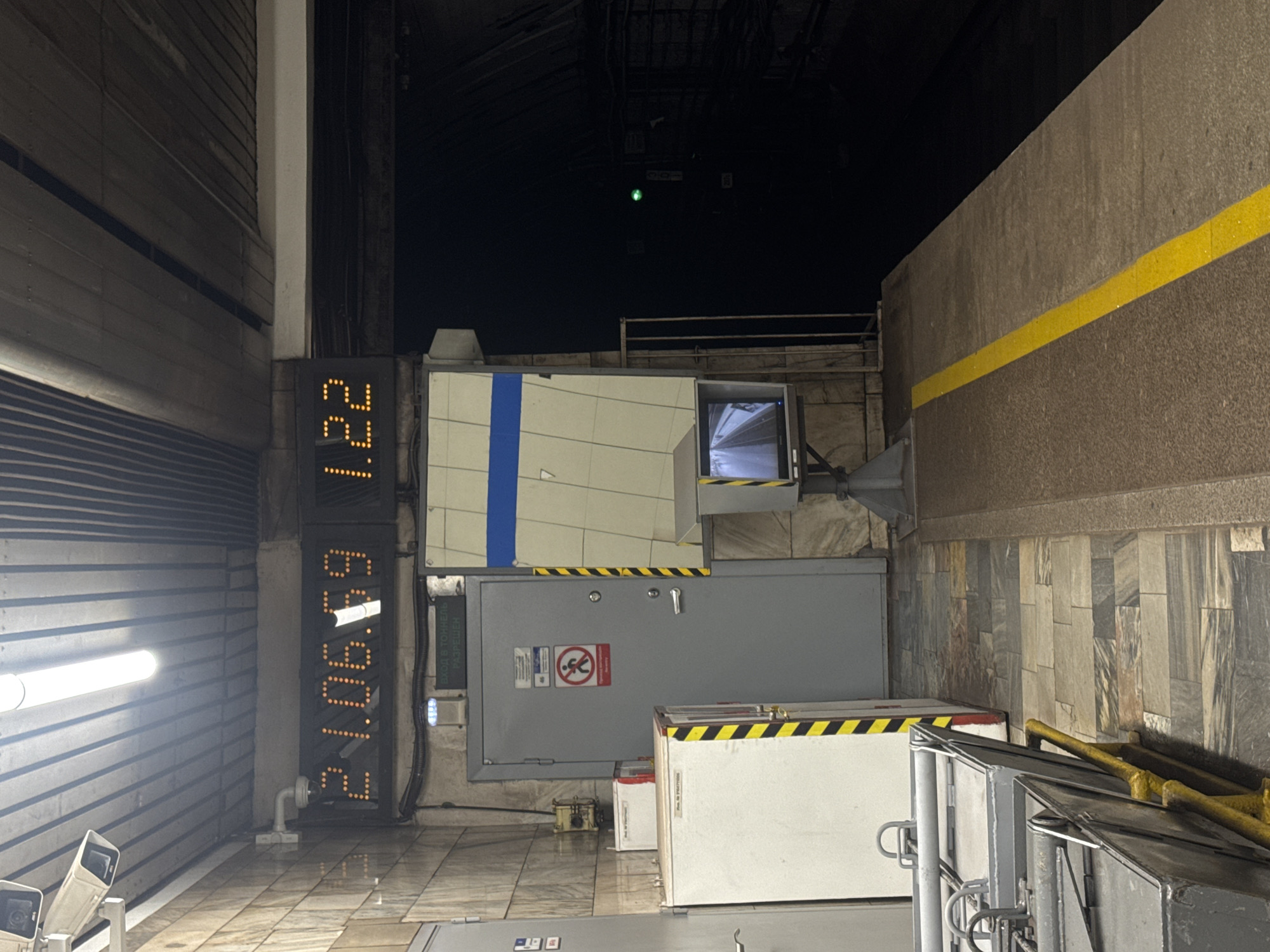 The image captures an indoor or covered outdoor scene, likely a passageway or tunnel entrance, at night. The setting appears to be industrial or utilitarian, with concrete walls and metal structures.
Prominently featured on the left is a large, vertical digital display showing bright orange numbers on a black background. The top part reads "221", and the bottom displays "699012". To the left of this display is a section of a corrugated metal wall, illuminated by a long, fluorescent light fixture. Two security cameras are visible at the base of the metal wall.
In the center of the image, a large grey door is set into a panel. Above the door, a smaller box-like structure with a screen is mounted. The screen displays a live camera feed showing a dimly lit area. The door has several signs attached, including a red circular sign with a diagonal line through a figure and a smaller rectangular sign with text.
To the right of the grey door, a white box with yellow and black hazard stripes is positioned. Behind this box, yellow railings are visible. The right side of the image shows a textured concrete wall with a bold yellow stripe running horizontally. The overall lighting suggests it is nighttime, and the environment appears to be dimly lit, with artificial lights illuminating the immediate area. The presence of security cameras and hazard markings indicates a functional and possibly restricted space.
Visible text on the rectangular sign next to the grey door reads: "ВХОД В ТОННЕЛЬ РАЗРЕШЕН" which translates from Russian to "ENTRANCE TO THE TUNNEL IS PERMITTED".