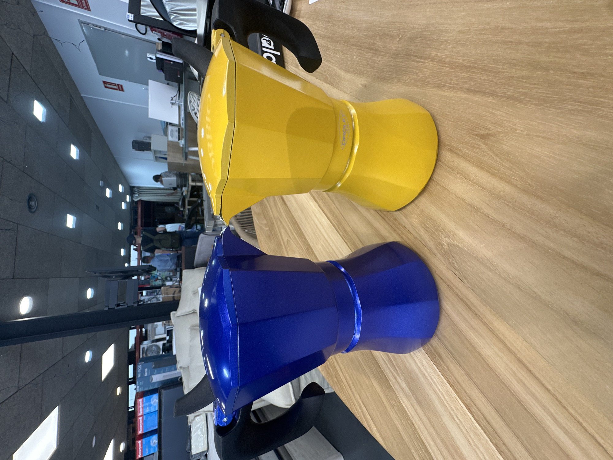Two vibrant Moka pots, one yellow and one blue, are prominently displayed on a light-colored wooden surface with a distinct grain. The image is oriented vertically, making the Moka pots appear to be lying on their sides. Both coffee makers feature a classic octagonal design with black handles. The yellow Moka pot has "D'ORO" subtly inscribed on its body and part of the "Bialetti" brand name visible on its black handle. The blue Moka pot has a metallic sheen.
The setting appears to be an indoor commercial space, possibly a showroom or store in Andorra la Vella, Andorra. The background is intentionally blurred, directing focus to the Moka pots. Visible in the soft-focus background are various elements indicating a retail or office environment. These include shelves, boxes (some clearly marked "Honeywell"), computer monitors, whiteboards, and diverse lighting fixtures like square and round ceiling lights. A concrete or stone-like wall is visible on the left side of the frame. Blurry figures of people can be seen in the distance, some seemingly working on laptops or browsing, suggesting typical daytime activity within the space. A red sign with "EXTINTOR" (fire extinguisher) and another small sign marked "EI" are also discernible on the back wall. The overall impression is that of products showcased in a modern retail or display area.