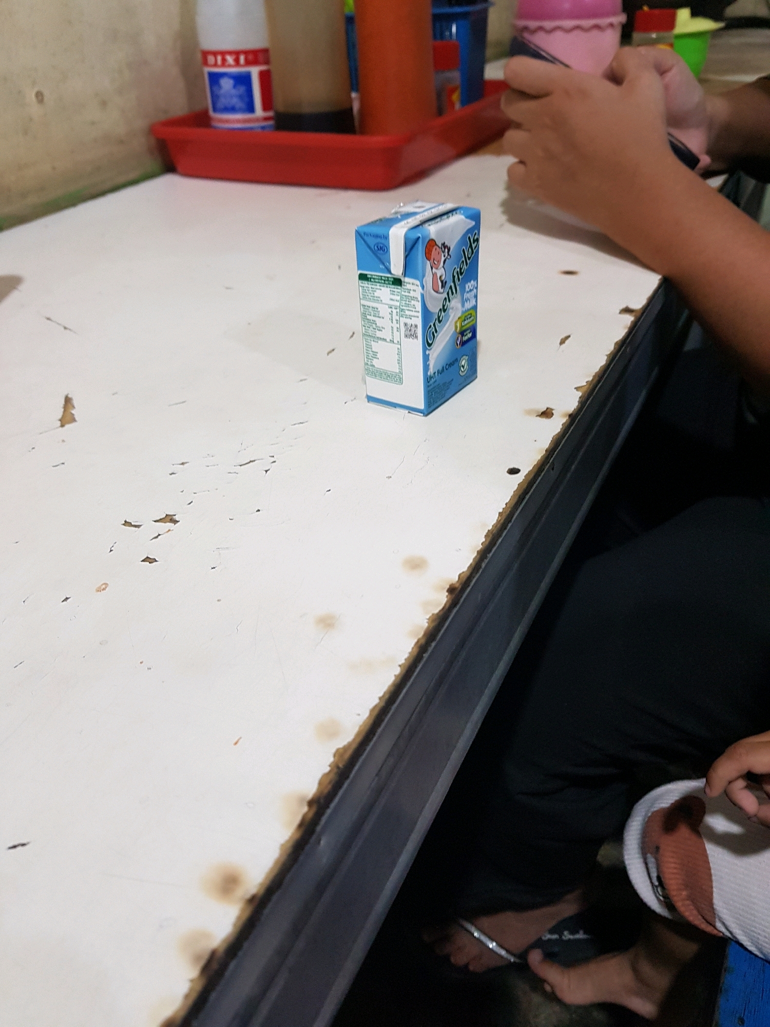 The image captures an indoor scene at what appears to be a casual eatery or home in Setiamekar, Indonesia. A white table with visible wear and stains occupies the foreground. On the table, a small carton of Greenfield's UHT Full Cream milk is positioned to the left. To the right, a person's hands are visible, holding a pink object, possibly a cup or container. In the background, various items are partially visible, including a red tray with a white and blue bottle, a red bottle, and a green container. The overall lighting suggests an indoor environment, potentially with ambient or artificial light sources. The presence of the milk carton and the casual setting suggests a simple meal or snack.