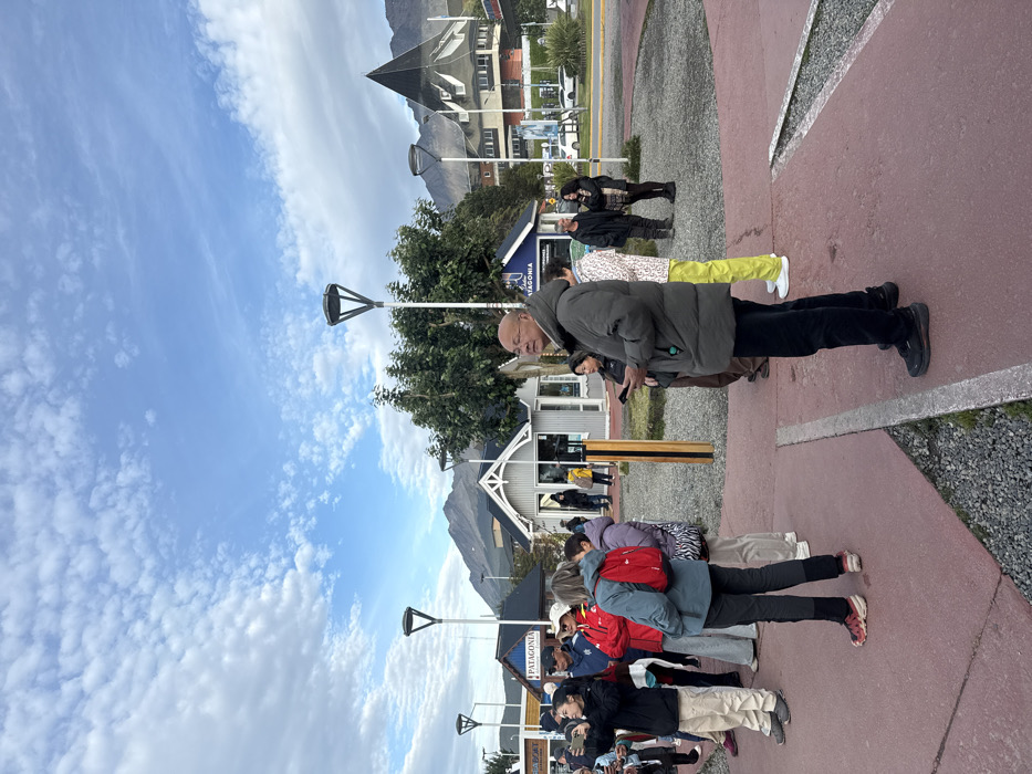 The image depicts an outdoor scene in Ushuaia, Argentina, specifically a pedestrian area or plaza adjacent to commercial buildings, with mountains in the background. The time is daytime, under a blue sky with scattered white cumulus clouds.
In the foreground, a red-paved surface with integrated strips of grey gravel and a concrete curb occupies the lower portion of the frame. Approximately 15-20 individuals, predominantly adults, are present in the scene. A bald East Asian male, wearing glasses and an olive-green padded parka over dark trousers, stands in the immediate foreground, facing right and looking forward, holding a dark smartphone in his left hand. To his left, a person wears a patterned jacket and bright yellow pants. Further left, a group of individuals observes the area; one woman in a dark jacket and light-colored pants holds a white smartphone as if capturing an image or video, and another woman in a blue-grey jacket with a red backpack faces away from the camera. Other individuals are distributed across the mid-ground, standing, observing, or interacting.
Modern streetlights with distinctive inverted-pyramid-shaped heads on white poles are spaced throughout the scene. Several small, light-colored commercial buildings with peaked roofs are visible in the mid-ground, one bearing "PATAGONIA" signage and another partially showing "BOAT". Green trees and bushes are interspersed between the streetlights and buildings. In the far background, a prominent multi-story building with a steep, dark, pointed roof is discernible, alongside other urban structures and a road with a white vehicle. Rugged, grey, treeless mountains dominate the horizon behind the urban landscape, characteristic of the region.
