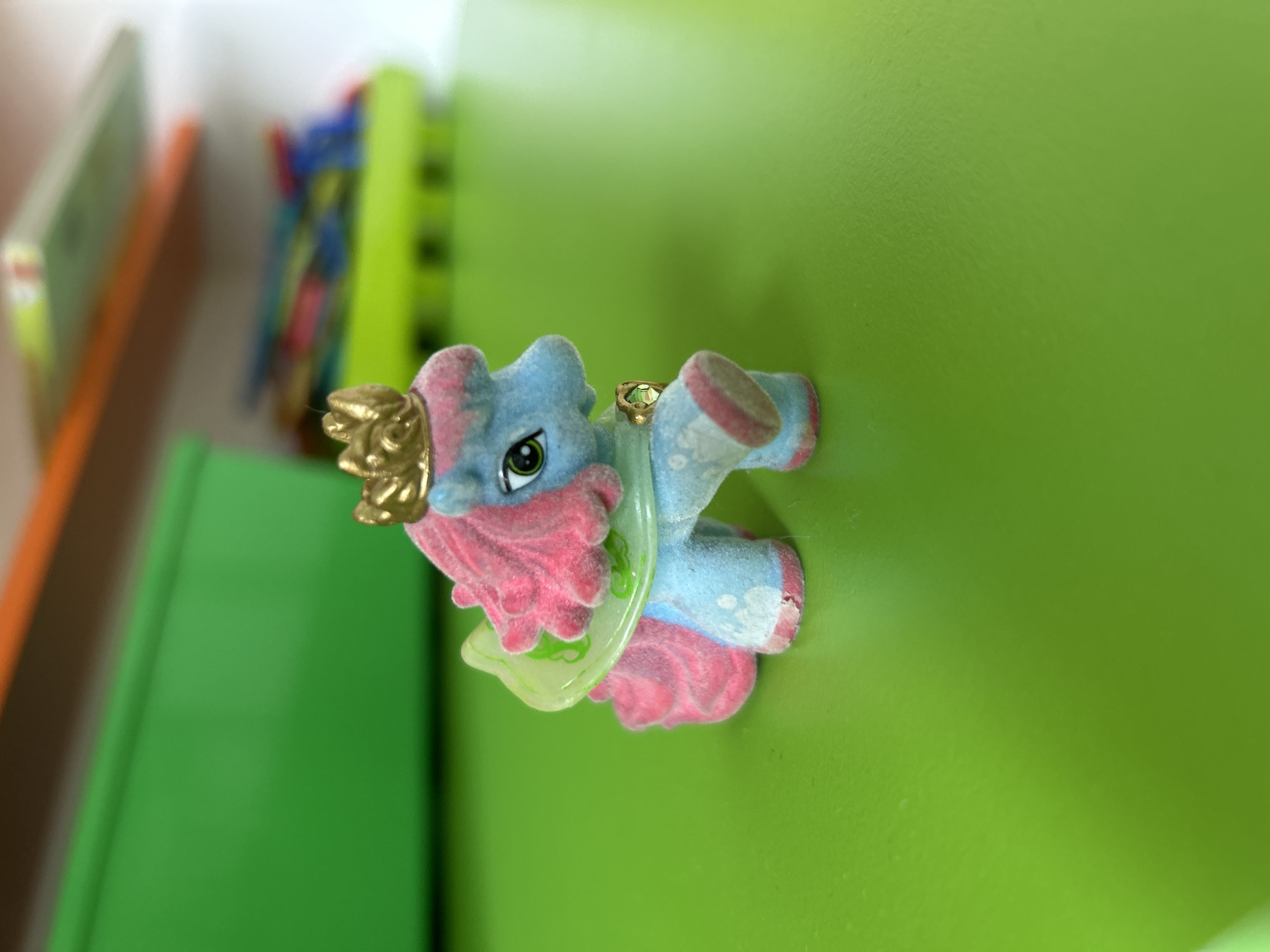 A fuzzy, blue toy horse with pink hair and a golden crown is positioned on a bright green, glossy surface. The horse has green eyes and a small, jeweled accessory around its neck. In the background, a blurred green bin and a colorful toy are visible on shelves. The scene is indoors and appears to be a child's play area. There is no information about the city of Khimki, Russia, in the image.