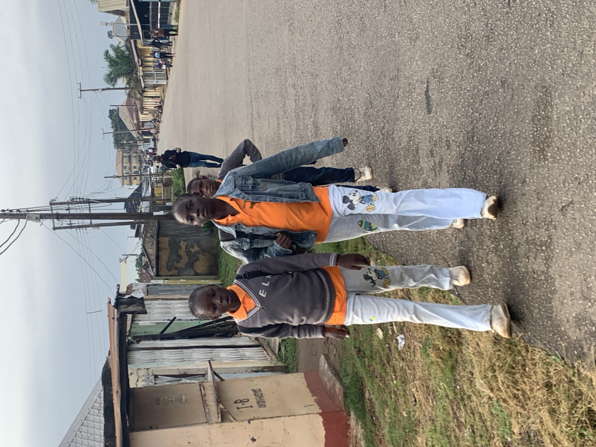 In Jos, Nigeria, three young individuals are walking along a paved street lined with low-rise buildings and structures. The sky is overcast. Two individuals are in the foreground, walking towards the viewer. The person on the left is wearing a grey v-neck sweater over an orange polo shirt and light-colored pants with cartoon characters printed on them. They have a backpack on. The person next to them is wearing an orange polo shirt and light-colored pants, also with cartoon prints, and a denim jacket. They are also carrying a backpack. A third person, further behind, is partially visible. In the background, other people are walking and standing, and a car is visible on the street. The buildings on either side of the street appear to be a mix of residential and commercial structures, some with corrugated metal roofs.