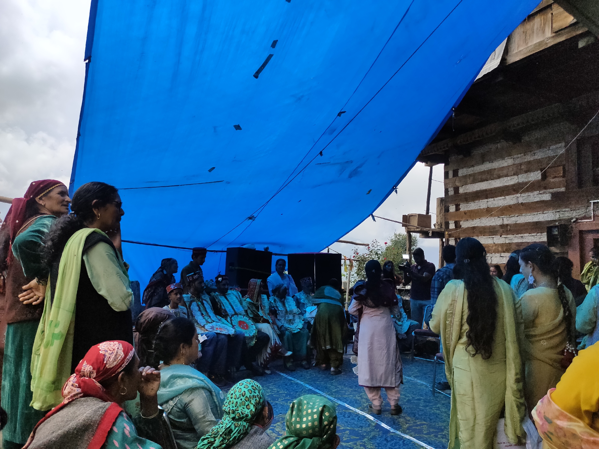 The image depicts an outdoor gathering under a large blue tarp, possibly in the village of Damadi, India. A group of people, predominantly women dressed in traditional Indian attire, are assembled. Some individuals are seated, while others are standing. The setting appears to be rural, with a rustic wooden building visible in the background. The overall atmosphere suggests a community event, perhaps a celebration or a meeting. The blue tarp, stretched overhead, serves as a makeshift roof, shielding the attendees from the elements. The cloudy sky hints at overcast weather.