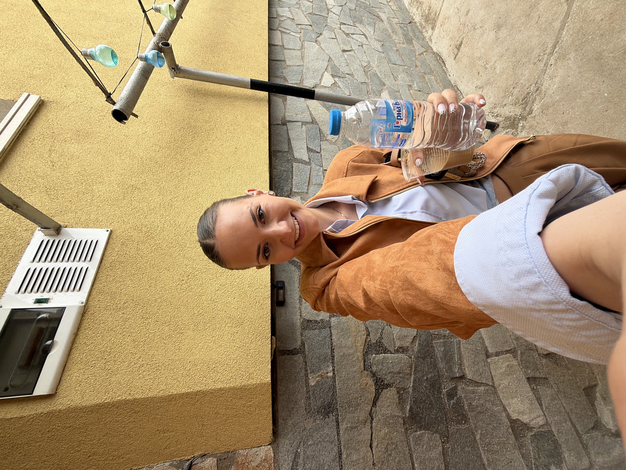A young woman is holding a water bottle, posing for a picture outside, likely in Buhovo, Bulgaria. She is wearing a tan jacket over a white top and a light blue shirt. The background shows a yellow wall with a ventilation unit and a metal structure with decorative light bulbs. The ground is paved with stone. The brand name on the water bottle appears to be "Горна Баня" (Gorna Banya).