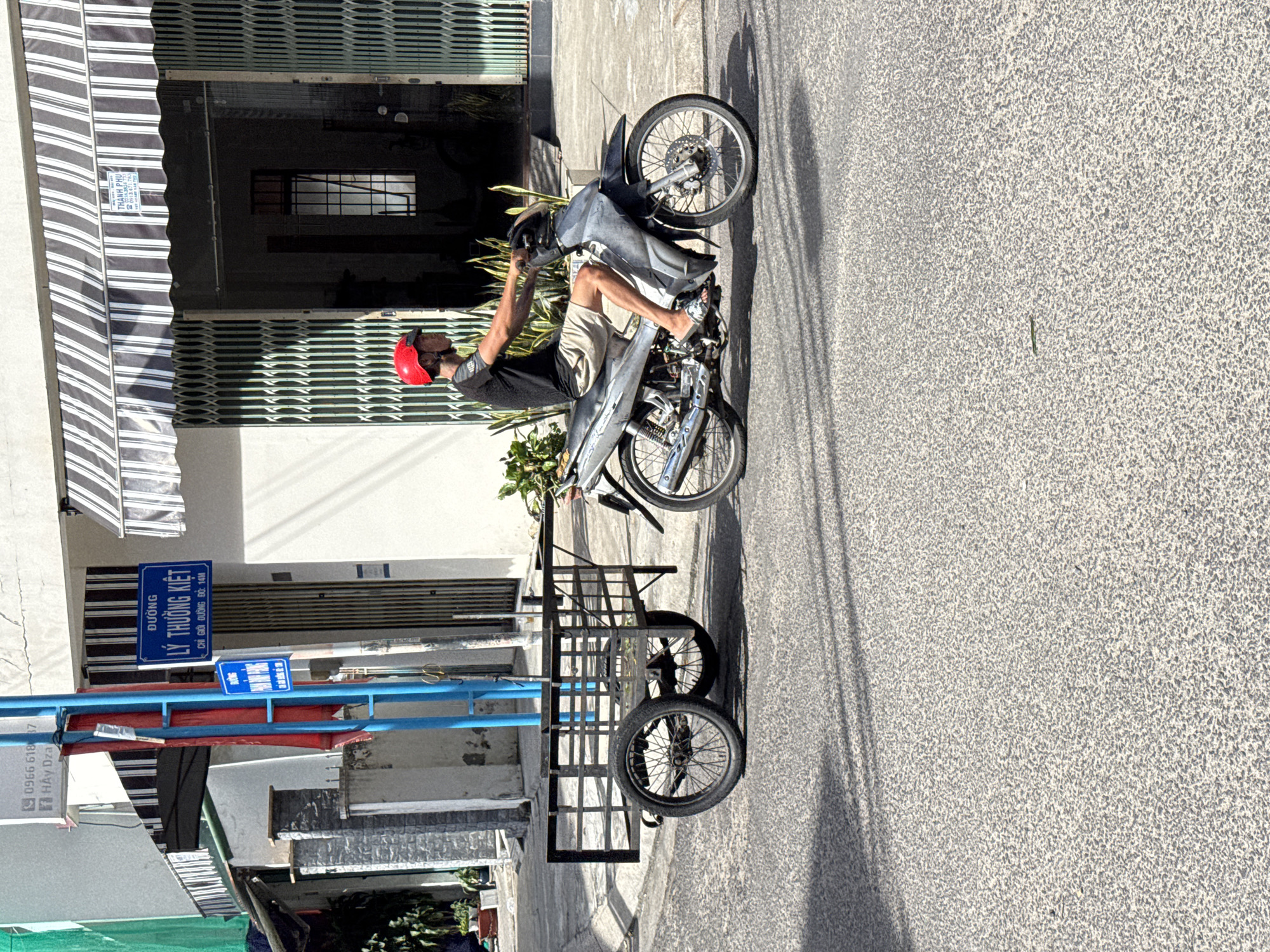 A man wearing a red helmet and a black t-shirt rides a silver motorcycle towing a metal cart. The cart has two wheels and a wire cage. The man is on a paved street in Nha Trang, Vietnam. There is a building with a striped awning in the background. A street sign in Vietnamese is visible on the left side of the image.