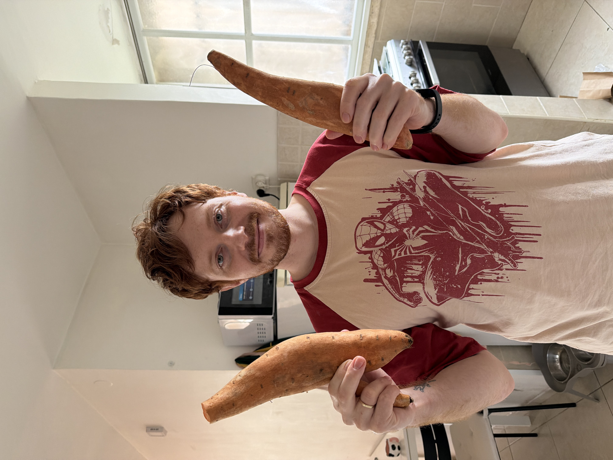 A male individual with reddish-brown curly hair, a light beard, and light-colored eyes stands centrally in a domestic kitchen, looking directly at the camera with a slight smile. He is wearing a light beige t-shirt with dark red raglan sleeves, featuring a large dark red graphic print of Spider-Man. A gold-colored ring is visible on his left ring finger, and a black fitness tracker is on his right wrist. A small blue tattoo is present on his left inner forearm. He holds a large, unpeeled sweet potato in each hand, angled upwards.
The kitchen features white-painted walls, with visible discoloration or water staining near the ceiling above a multi-pane window on the right side. A white electrical outlet is on the wall to the left. The floor is covered with light-colored square tiles. Appliances include a stainless steel gas range/oven on the right and a white microwave oven positioned above a white refrigerator on the left. A white table with a dark chair is partially visible on the far left, along with a small black and white decorative object. Two metal pet bowls are located on a stand on the floor near the individual's left arm. A brown paper bag and some plastic are on the floor near the oven.
The scene depicts an indoor domestic setting. While consistent with residential interiors found in Tel-Aviv, Israel, no specific geographical markers are present in the image to identify an exact location within the city.