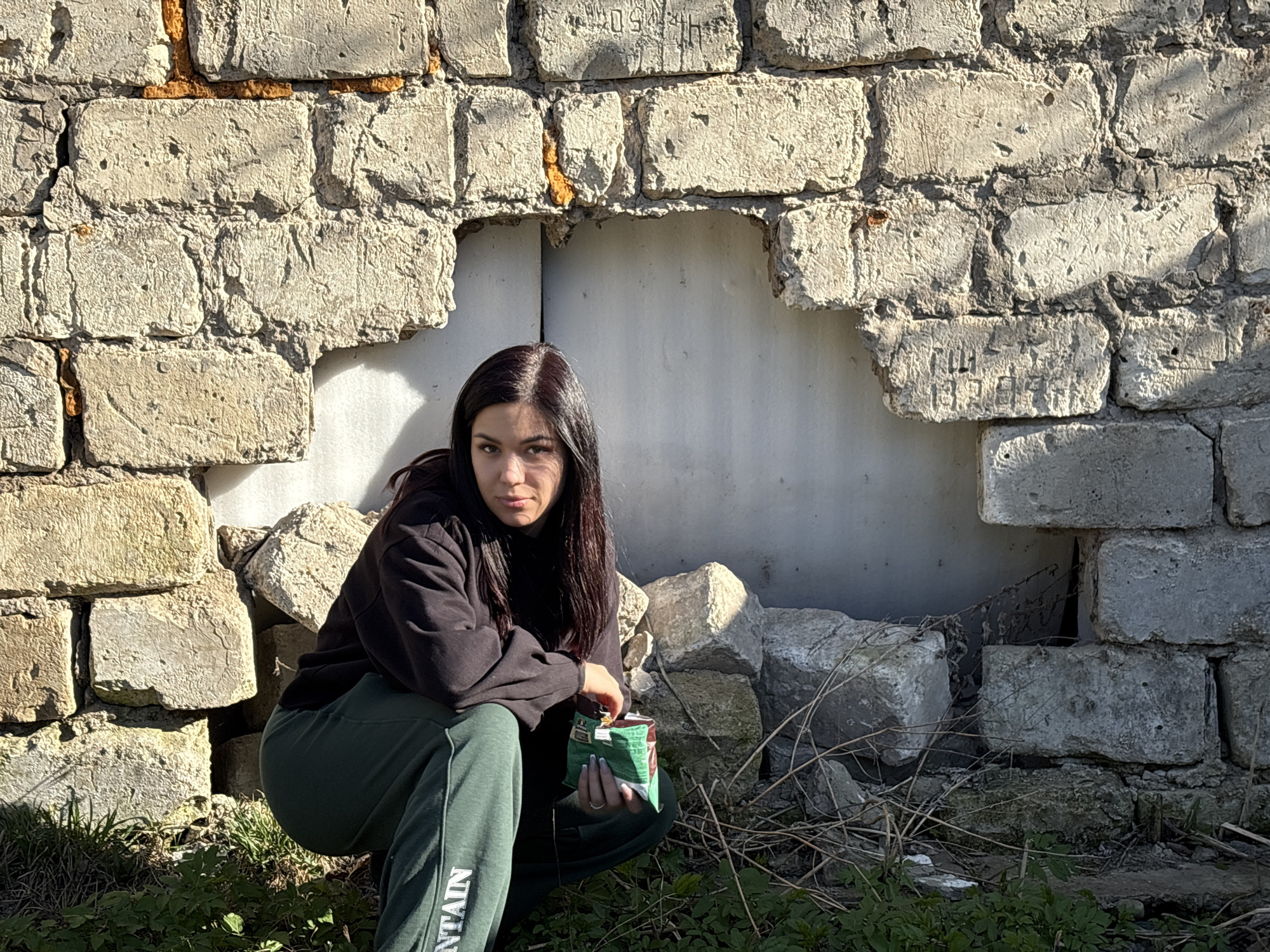 A young woman with long, dark hair is crouching in front of a dilapidated brick wall, holding a green and brown bag of chips. She is wearing a black hoodie and green sweatpants with the word "MOUNTAIN" printed vertically on the left leg. The wall has a large hole in it, revealing a light-colored surface behind. Debris from the wall is scattered around her on the ground, mixed with some weeds and dry vegetation. The setting appears to be urban, possibly an abandoned or neglected area. In the brick, "1954" is visible.
The location is Азинский, Russia.