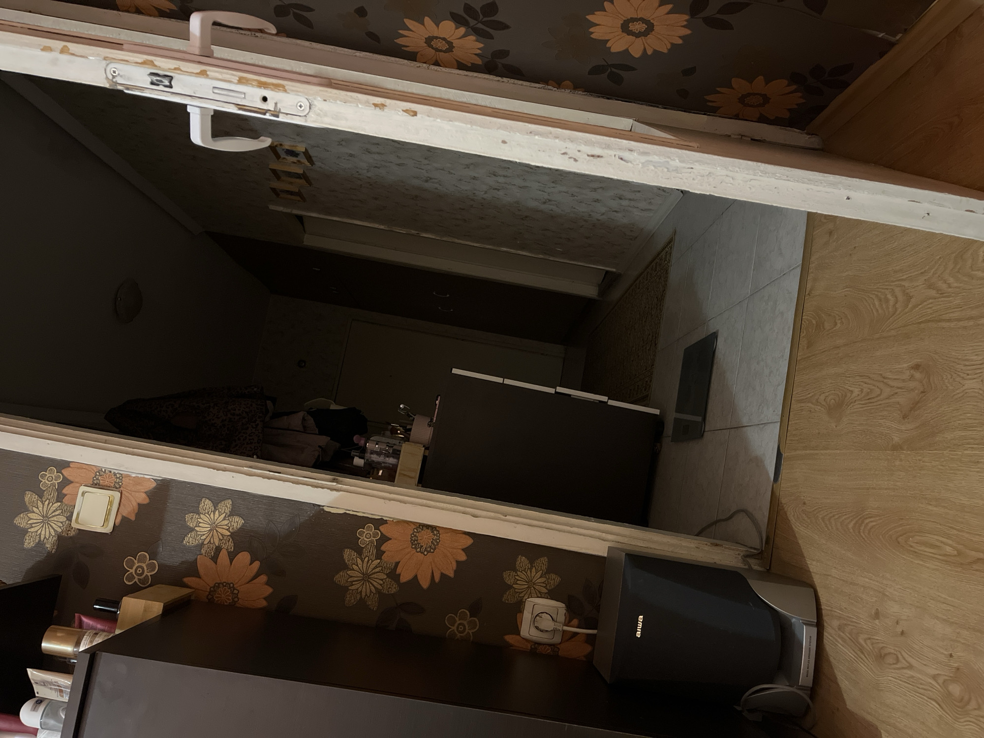 The image depicts an interior hallway, likely in a residential building, possibly in Plovdiv, Bulgaria. The hallway features a dark brown floral wallpaper on the right wall and a lighter, textured wallpaper on the left. A door frame, showing signs of wear and peeling paint, dominates the right side of the image, suggesting it is open.
Inside the hallway, on the left, a dark brown cabinet is visible, with some toiletries and makeup items placed on a shelf above it. Below the shelf, a black Aiwa speaker system is positioned on the floor, with a white electrical plug connected to a wall socket above it. The floor appears to be a laminate wood-grain pattern.
Further into the hallway, a doorway leads to another room. A light-colored rug lies on the floor. On the far wall, several framed pictures or objects are mounted. The lighting in the hallway is dim, casting shadows. The overall impression is one of a functional, yet somewhat cluttered, domestic space.