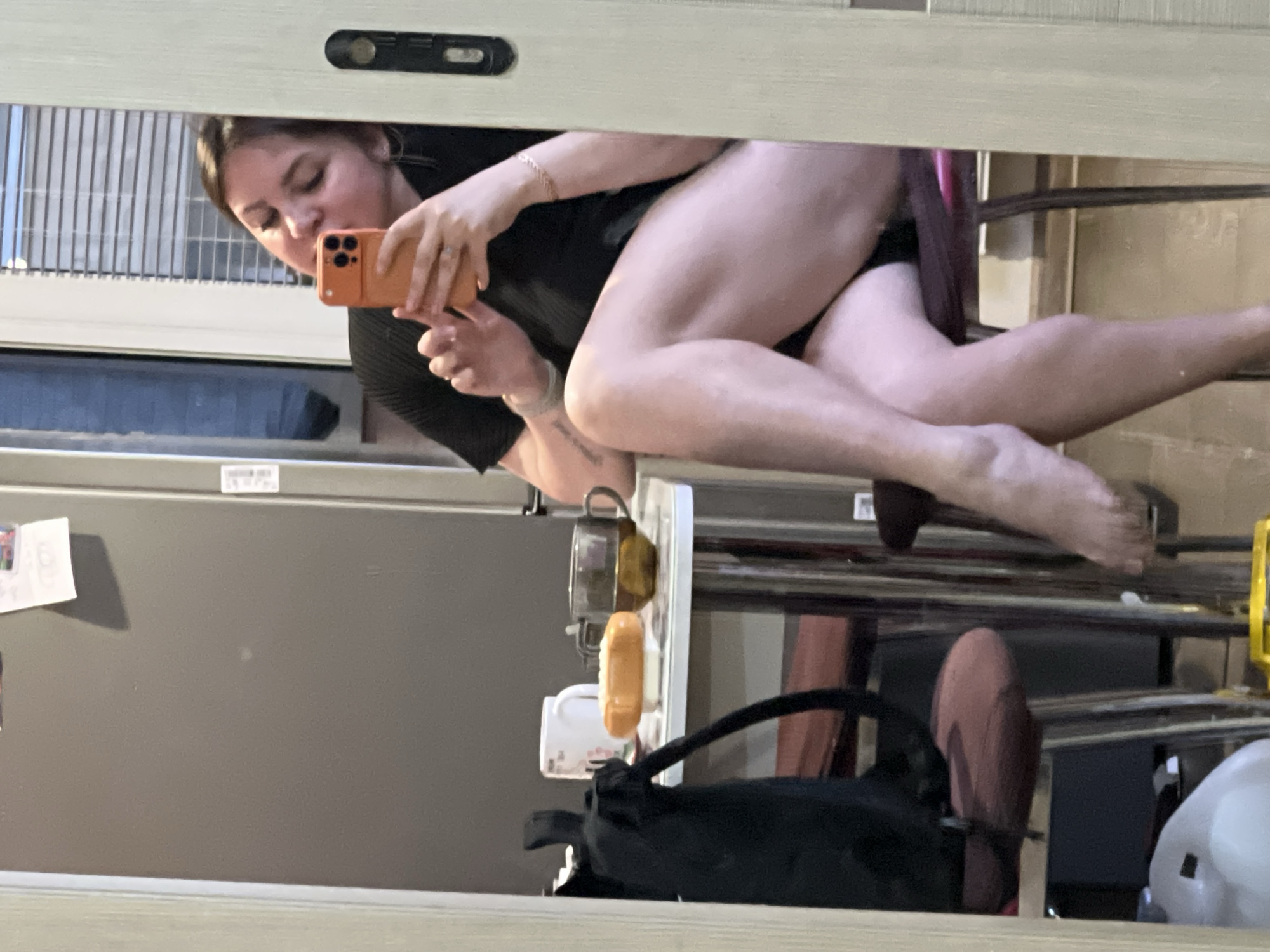 A woman is taking a selfie in a mirror. She is seated on a chair and holding an orange cell phone. The reflection shows a room with a window, a refrigerator, and some items on a shelf. The mirror frame is light-colored. There is no information to confirm it is in the city of Noviy, Russia.