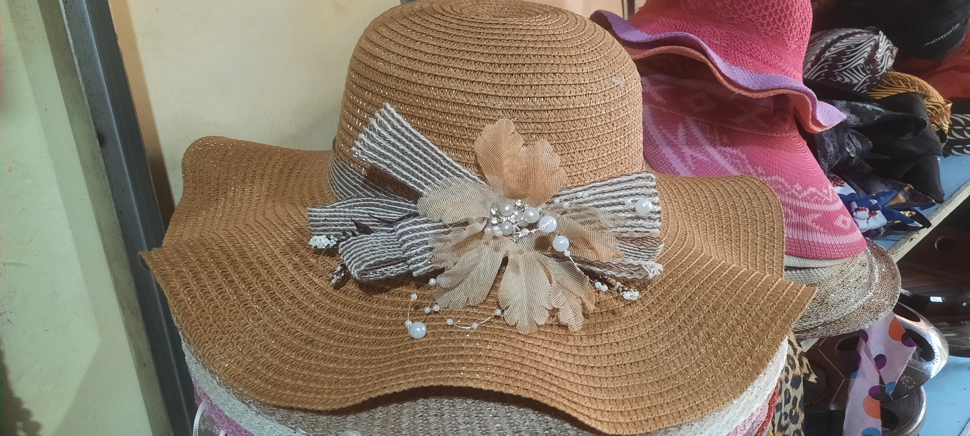 A close-up, high-angle, full shot shows a woven straw sun hat with a wide brim, decorated with a bow and faux pearls. The hat is positioned in the foreground, obscuring other items beneath it, including what appear to be other hats and scarves. The hat's natural straw color is a warm tan, and the brim is gently curved. A decorative element featuring a striped ribbon, sheer fabric petals in a muted peach color, and small white beads and faux pearls is centered on the front of the hat. The background is blurred, but shows other hats, including a bright pink one with a ruffled brim, and some patterned fabrics. The scene suggests a retail environment, possibly a stall or shop selling accessories. The lighting is even, highlighting the textures of the hat and decorations. The image is taken in Blitar, Indonesia.