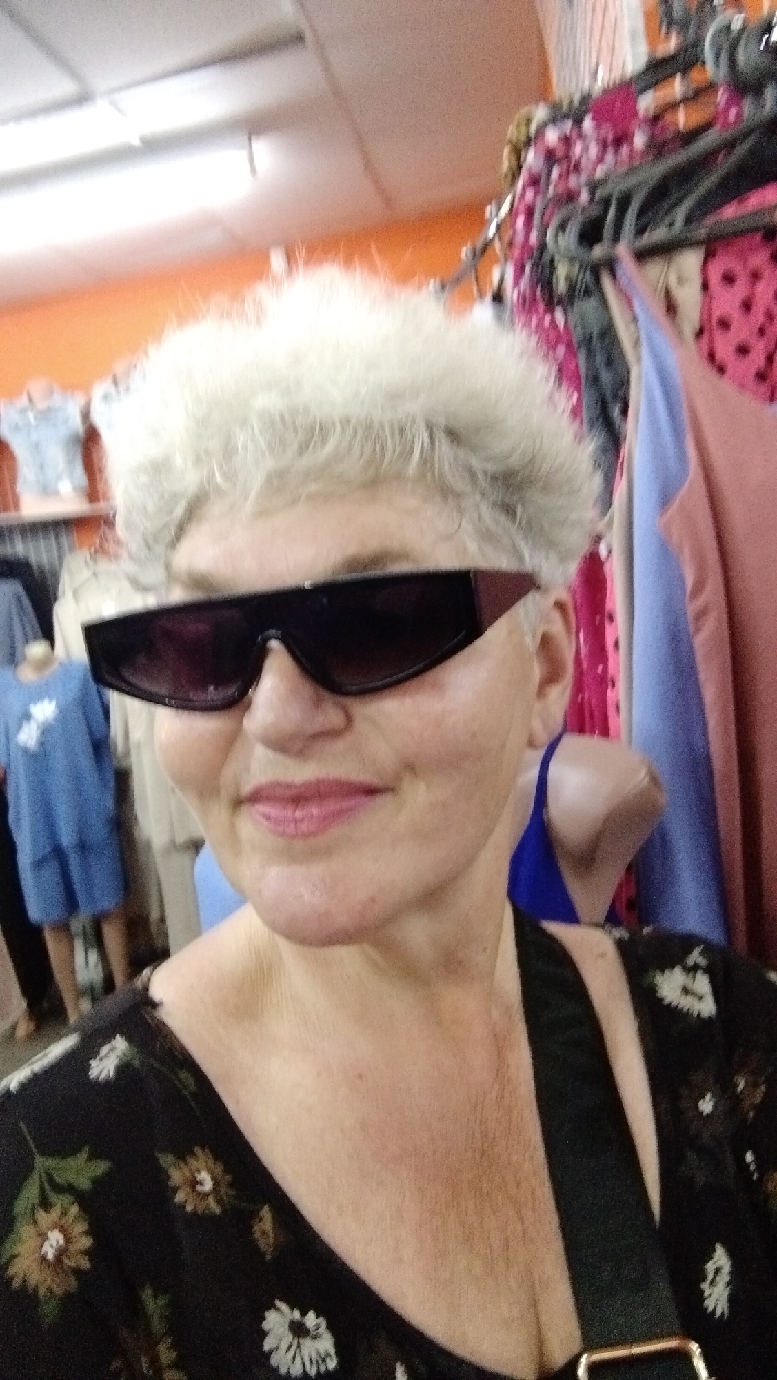A middle-aged woman with short, white, fluffy hair and sunglasses is posing for a photo indoors. She is wearing a black top with a floral pattern and a black strap across her chest. In the background, racks of colorful clothing are visible, suggesting the location is a store. The woman is looking directly at the camera with a slight smile. The lighting in the room is bright, likely from overhead fluorescent lights, casting a warm glow on the scene.